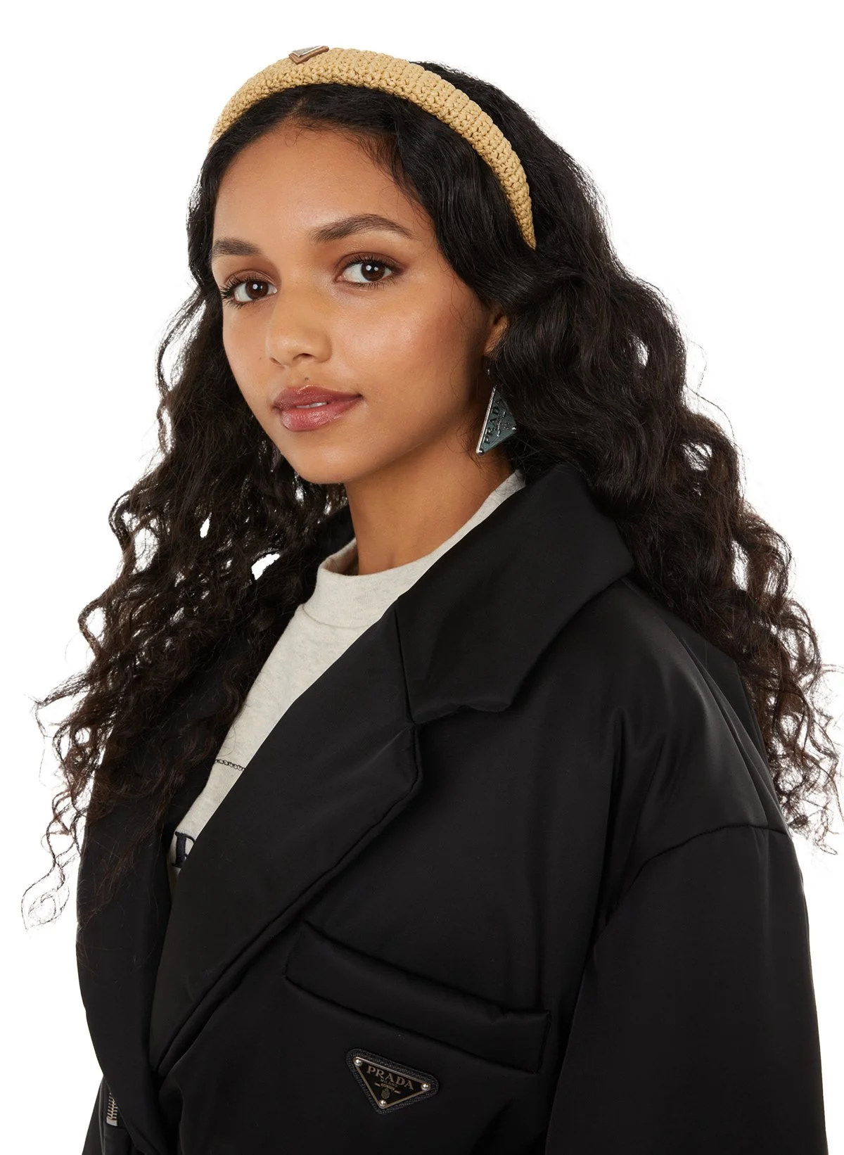 Jeune femme aux cheveux bouclés portant un headband beige, une veste noire, un t-shirt blanc, une boucle d'oreille triangulaire, avec un fond blanc.
