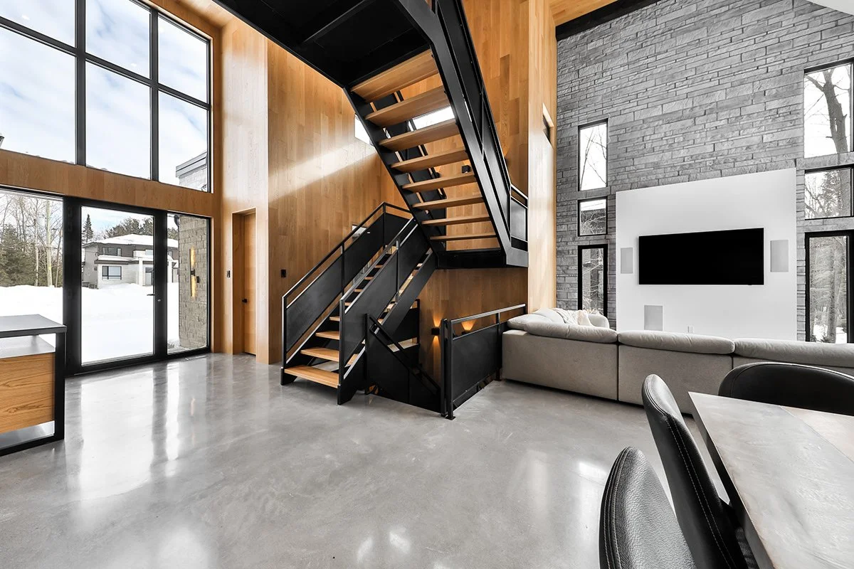 Intérieur moderne d'une maison avec escalier en bois et métal, murs en pierre et bois, grand séjour avec canapé gris et télévision sur le mur, fenêtres donnant sur un extérieur enneigé.