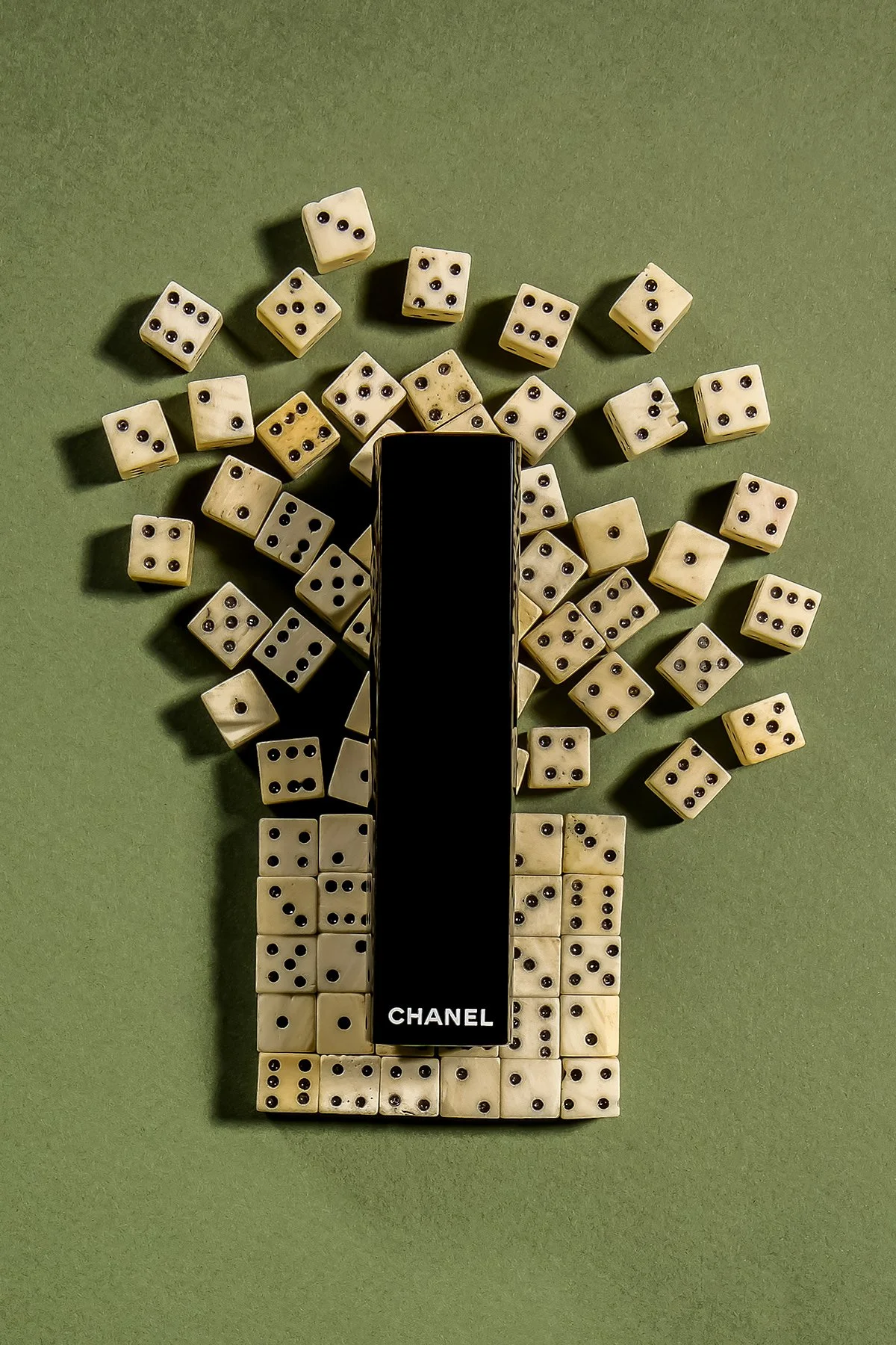 Une composition de dés blancs de différentes tailles autour d'une boîte noire portant la marque Chanel, le tout sur un fond vert.