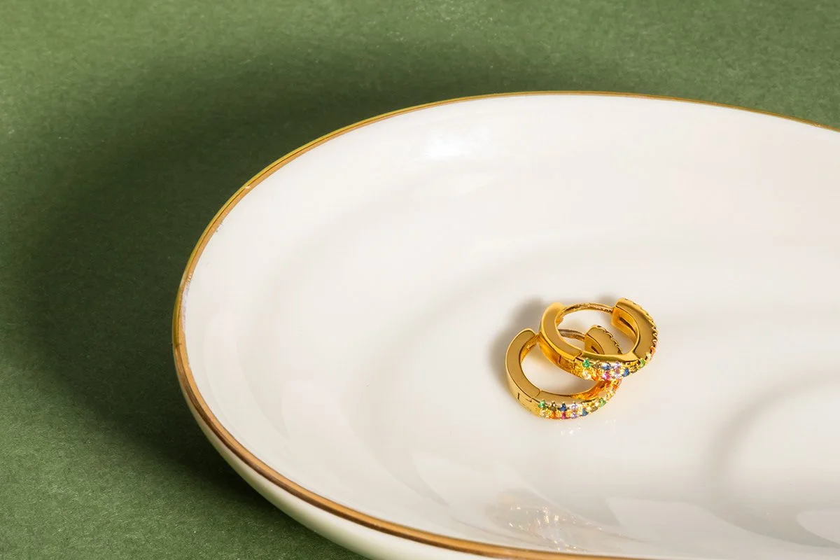 Deux bagues en or avec des pierres colorées, posées dans une assiette blanche avec une bordure dorée, sur un fond vert.