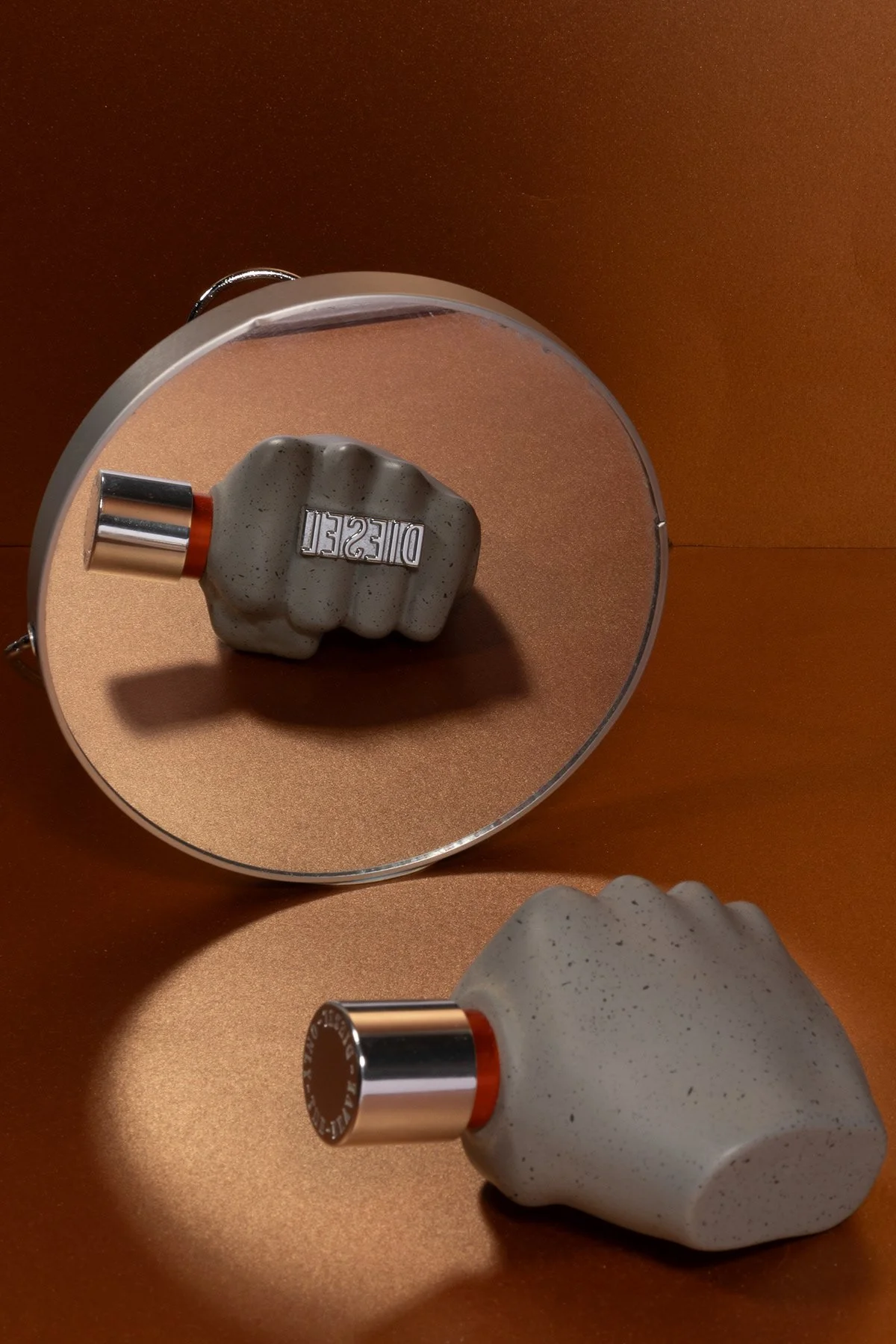 Une bouteille de parfum en forme de poing, posée sur un miroir rond sur une surface brun orangé.