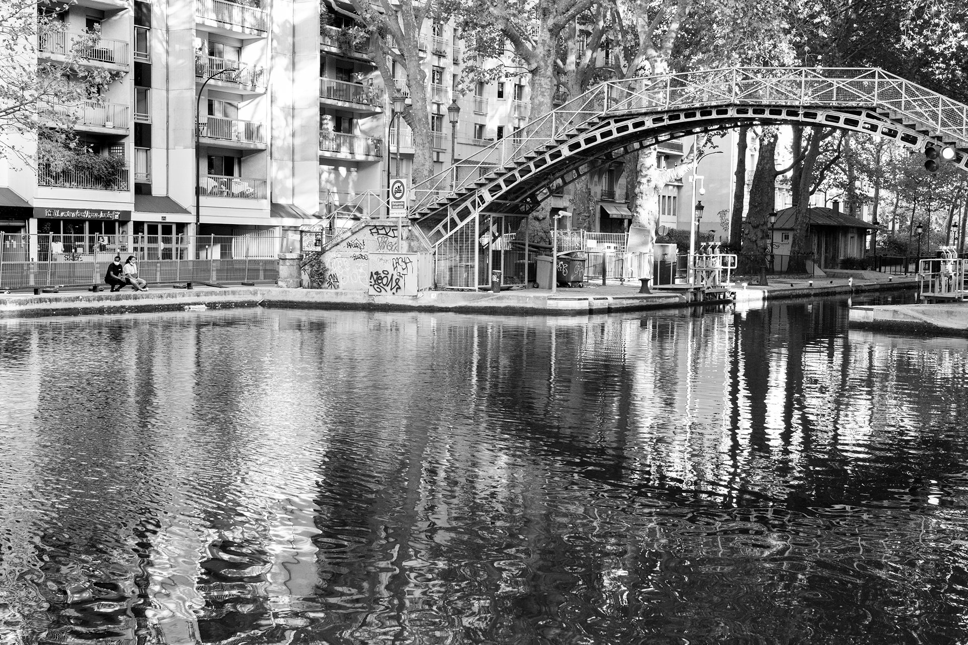 Une scène urbaine en noir et blanc avec un pont métallique au-dessus d'un canal, entouré de bâtiments résidentiels et d'arbres. Deux personnes sont assises sur un banc à gauche.