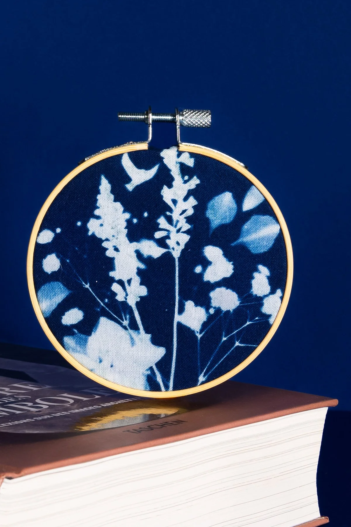 Un cercle de broderie montrant un motif floral de feuilles en blanc et bleu sur fond bleu foncé, posé sur un livre