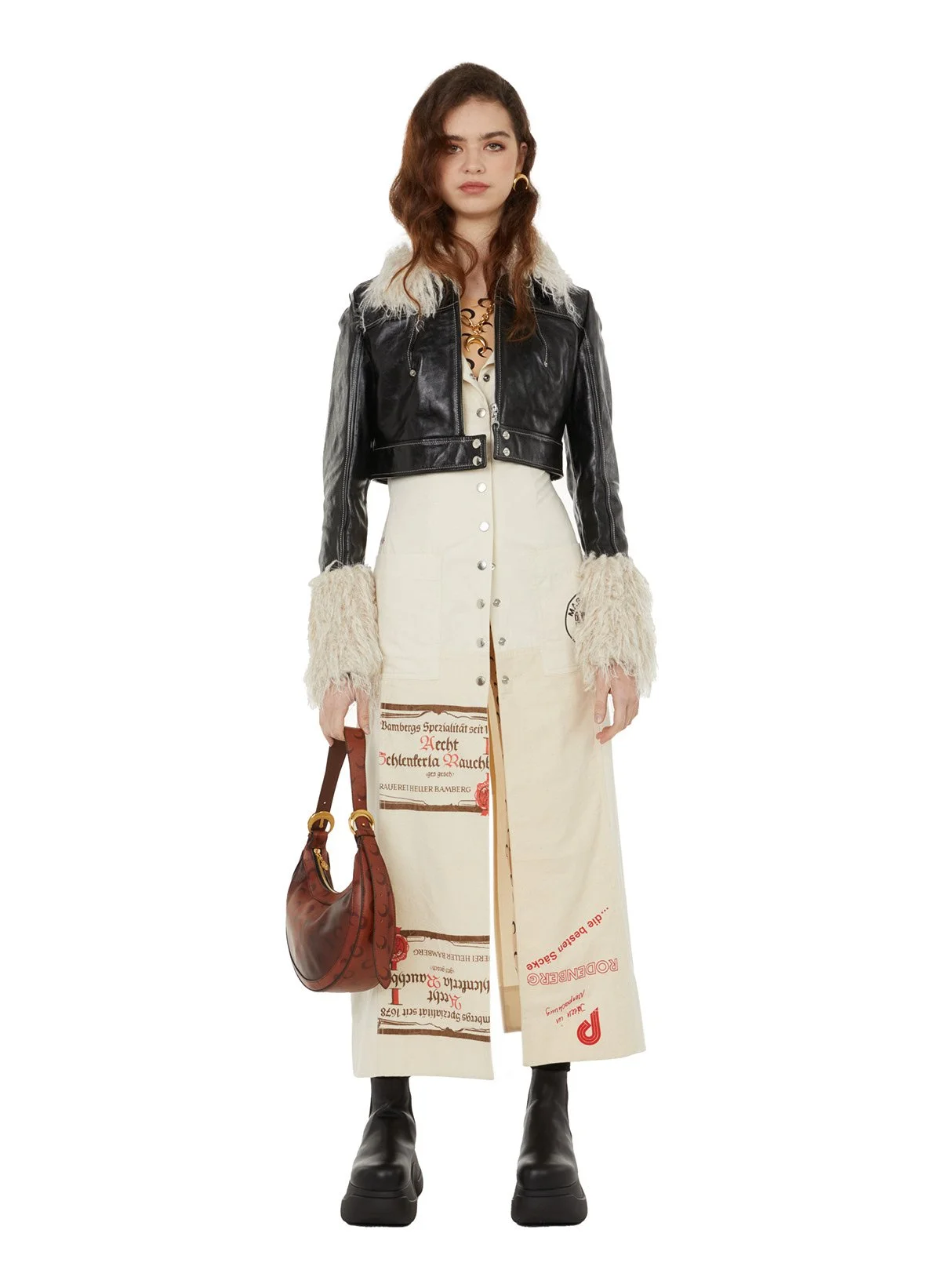 Une jeune femme porte une veste en cuir avec une doublure en laine, une robe longue à imprimés, des bottes noires, et tient un sac en cuir marron. Elle a des boucles d'oreilles dorées et des cheveux bruns ondulés.