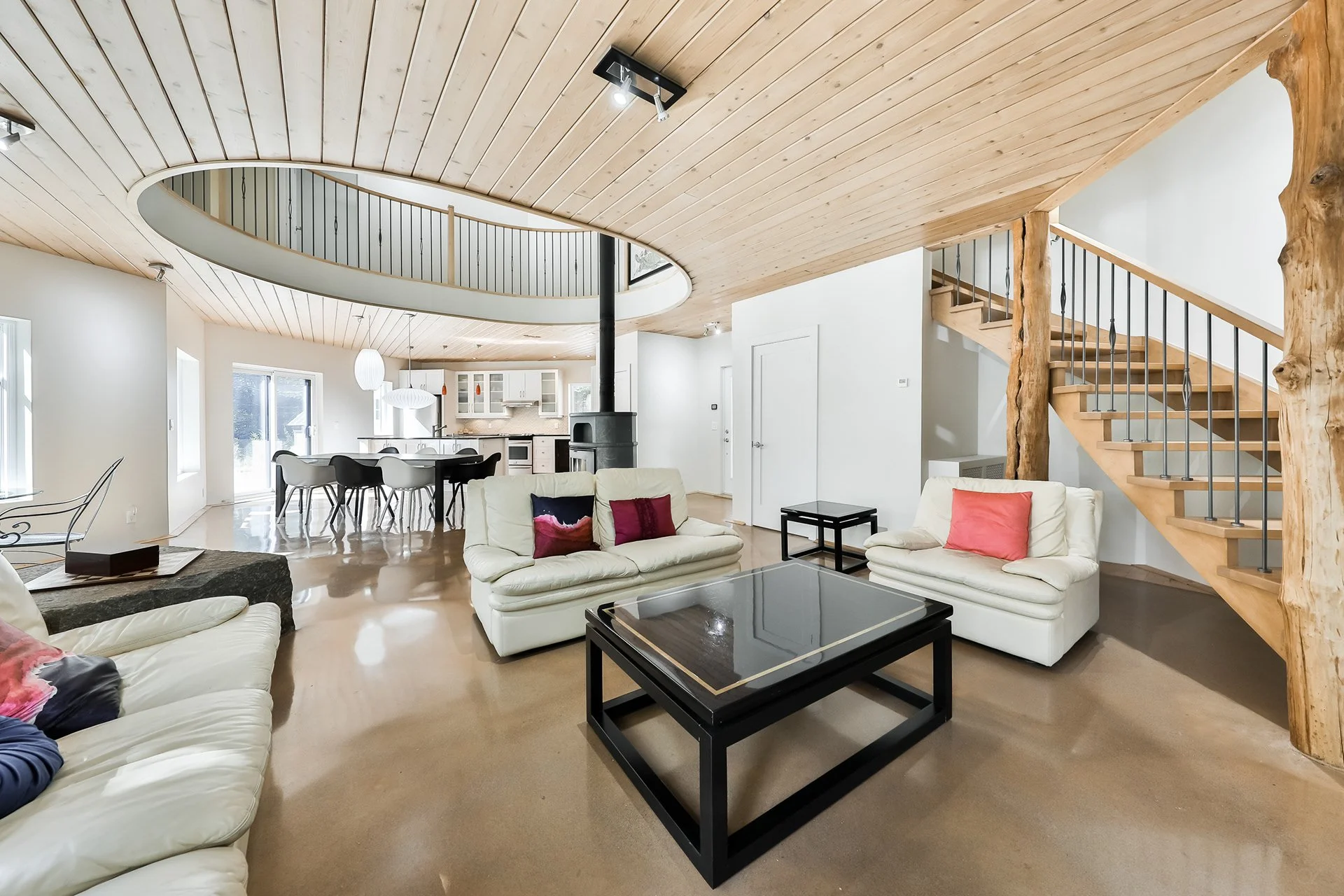 Salon avec canapés crème, coussins colorés, table basse noire, escalier en bois avec poteaux en bois naturel, plafond en bois, espace lumineux avec murs blancs et cuisine ouverte en arrière-plan