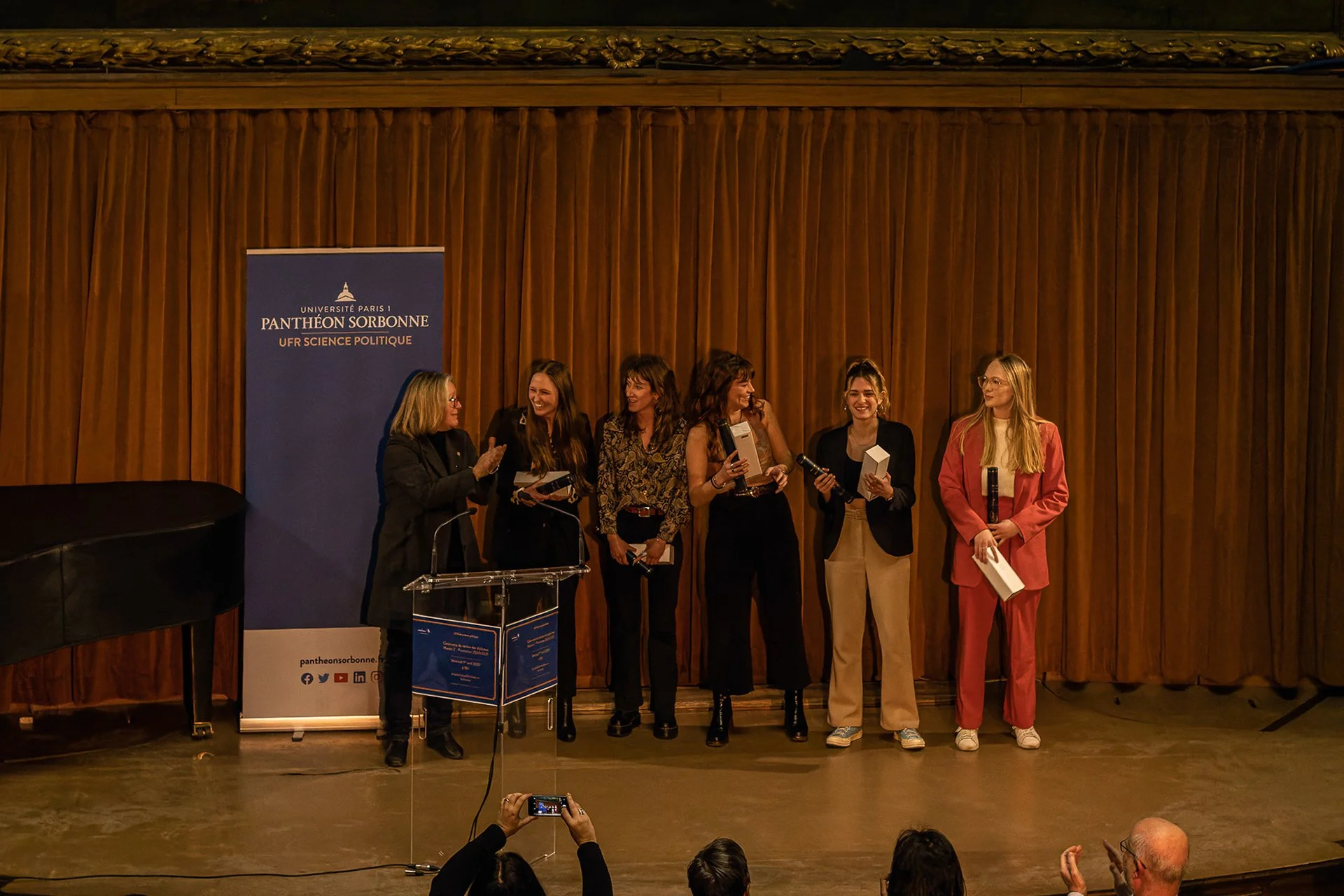 Six women standing on stage in front of a wooden background at an award ceremony. One woman is speaking at a podium with a Panthéon Sorbonne banner beside her. The other women are holding awards and smiling.