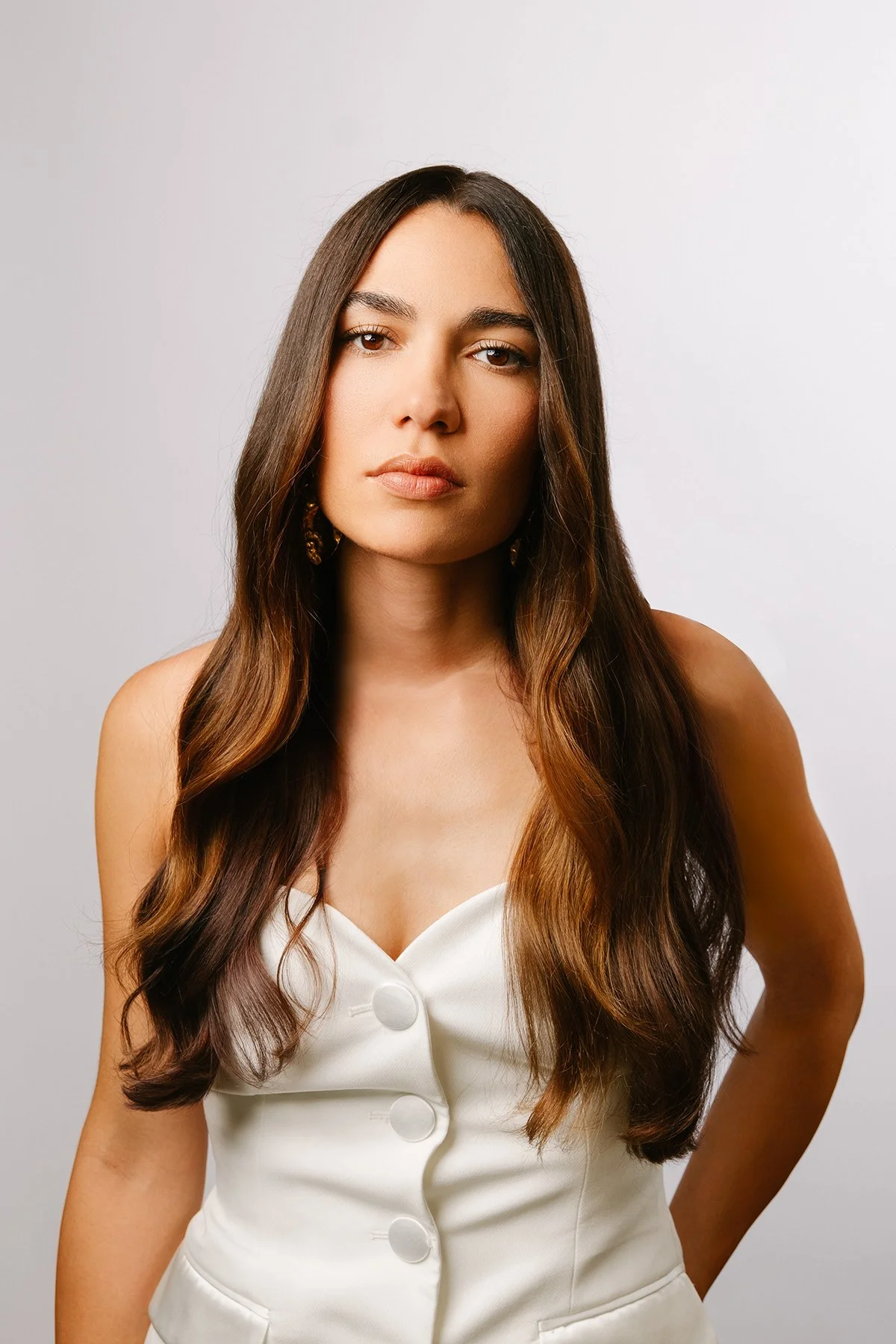 Portrait d'une jeune femme aux longs cheveux bruns portant une robe blanche à boutons, posant devant un fond neutre.