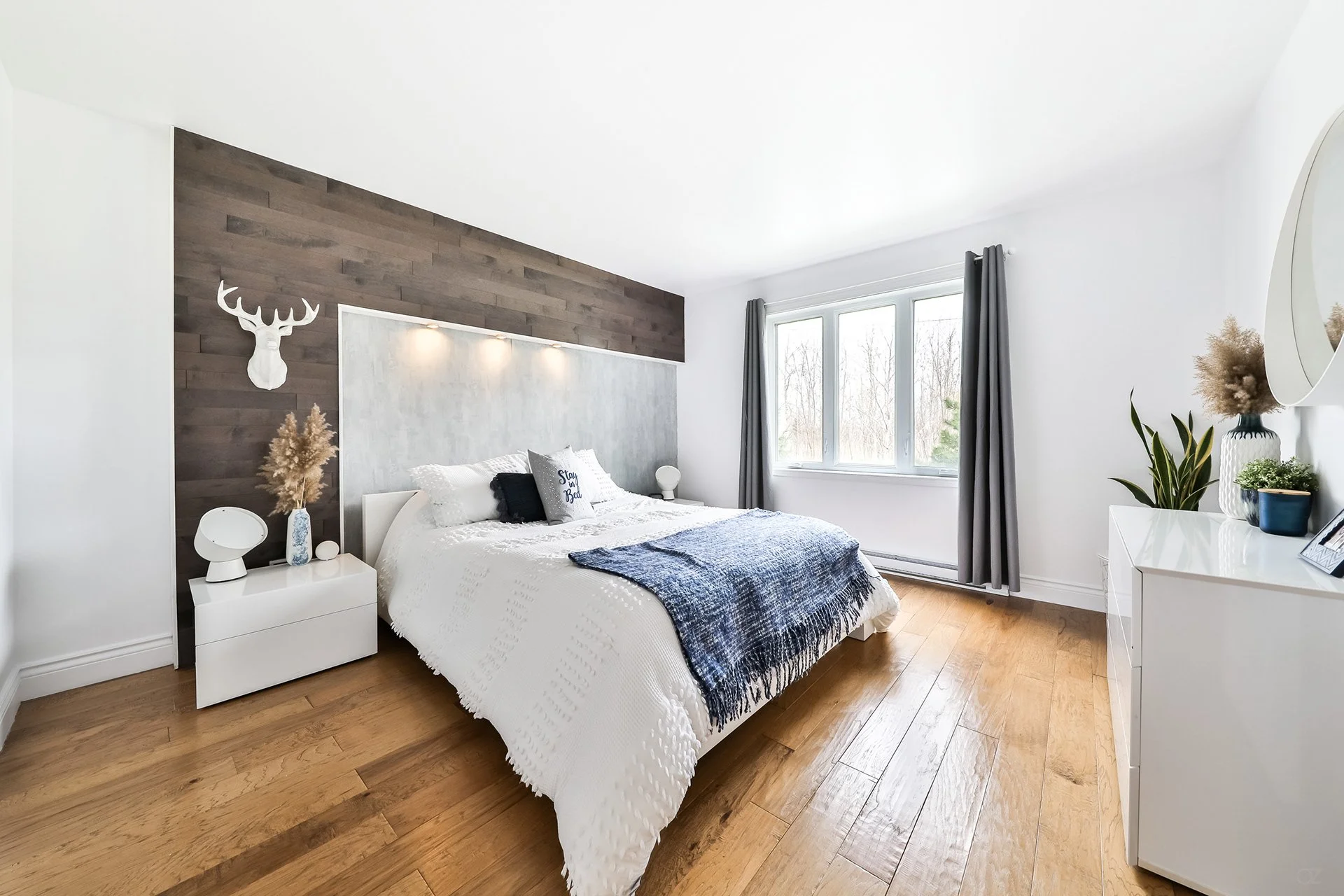 Chambre moderne avec un lit blanc, une tête de lit en bois foncé, une décoration de cerf en haut du mur, et des objets décoratifs sur une commode blanche, ainsi qu'un grand mur de fenêtres avec des rideaux gris.