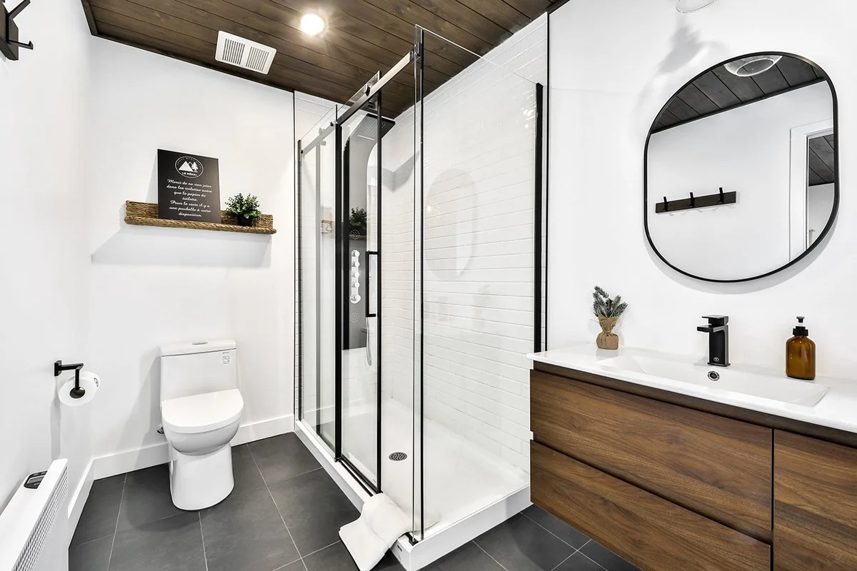 Salle de bain moderne avec douche en verre, miroir rond, meuble en bois, lavabo, et toilettes dans une décoration minimaliste.