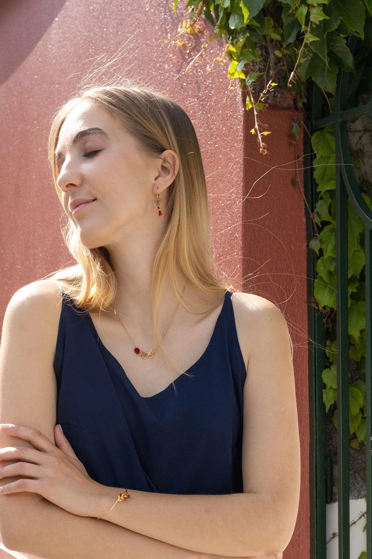 Une femme souriante portant des bijoux en or, une robe bleue, avec une coiffure aux cheveux longs, se tenant devant un mur rose avec de la végétation verte.