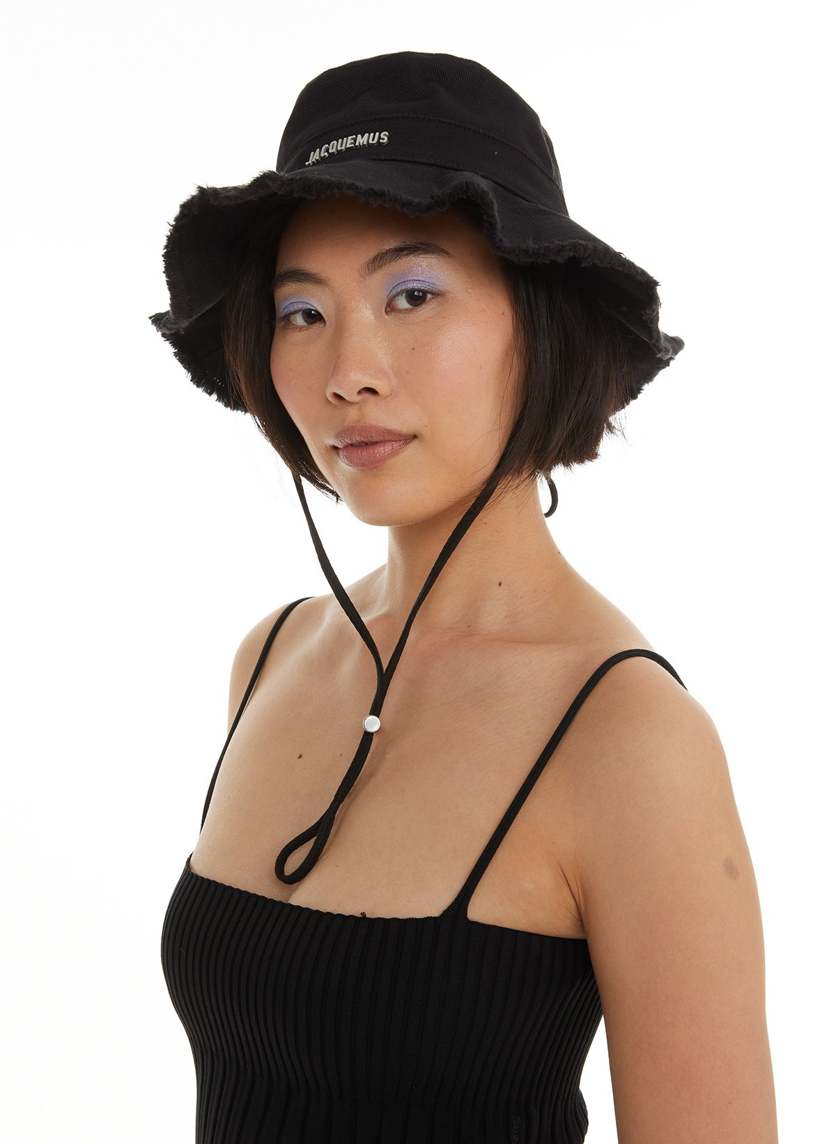 Femme portant un chapeau noir avec un logo blanc, vêtue d'un haut noir à bretelles, contre un fond blanc.