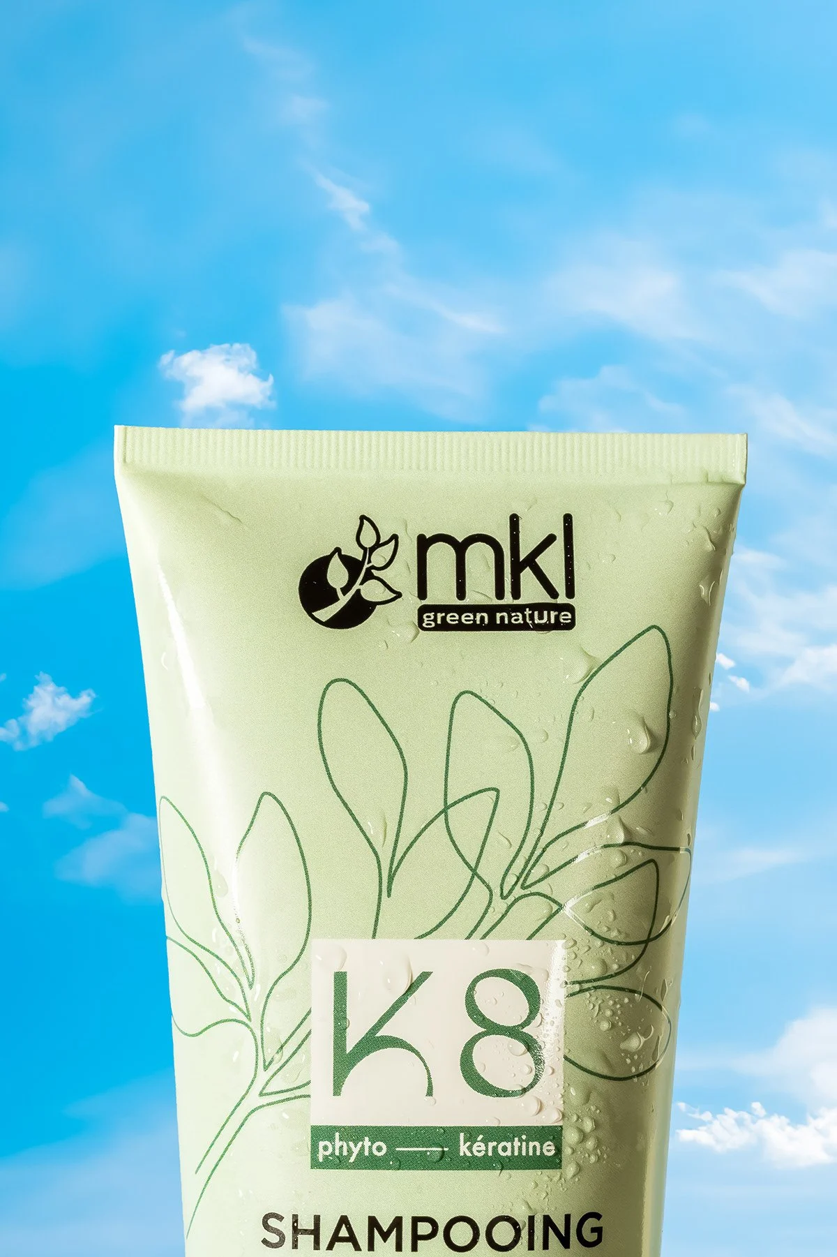 Une bouteille de shampoing vert avec des illustrations de feuilles, étiquetée 'K8 phyto kératine', contre un ciel bleu avec quelques nuages.
