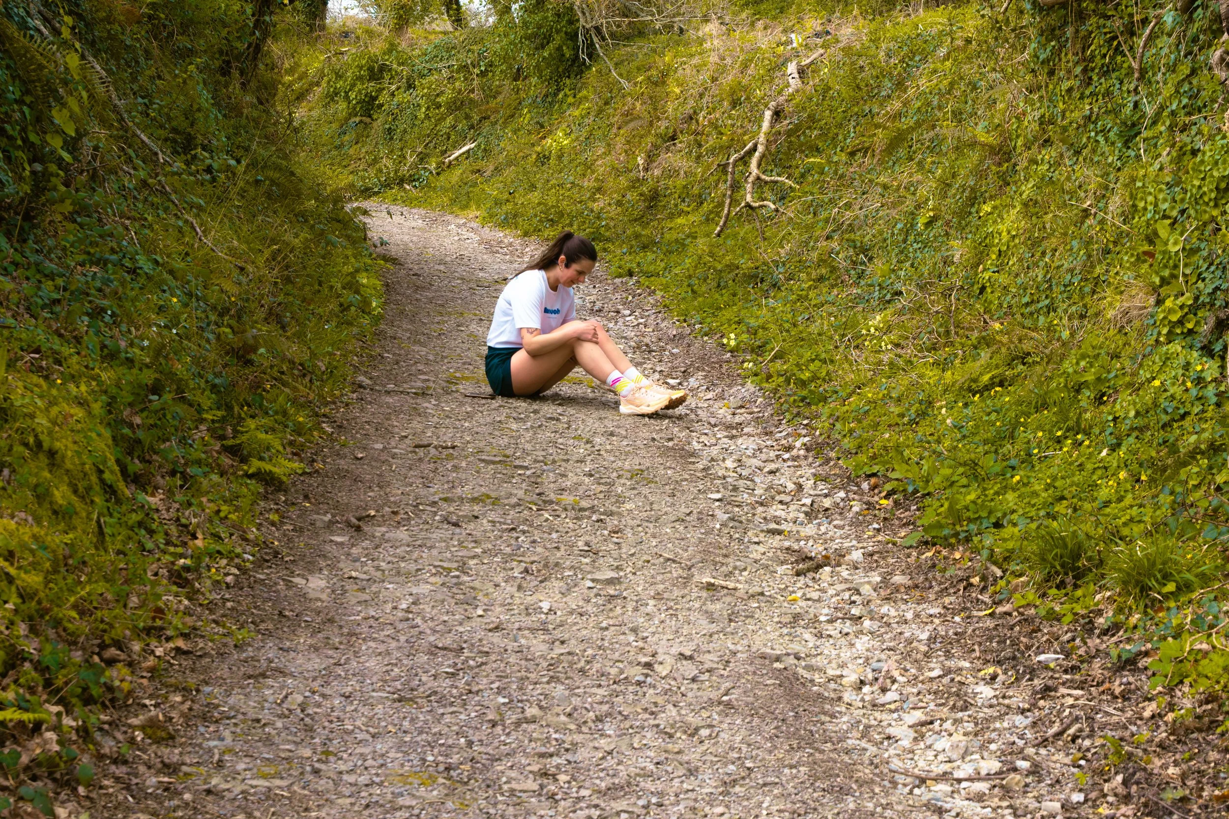 Une jeune femme assise sur un sentier de campagne entouré de végétation verte dense.