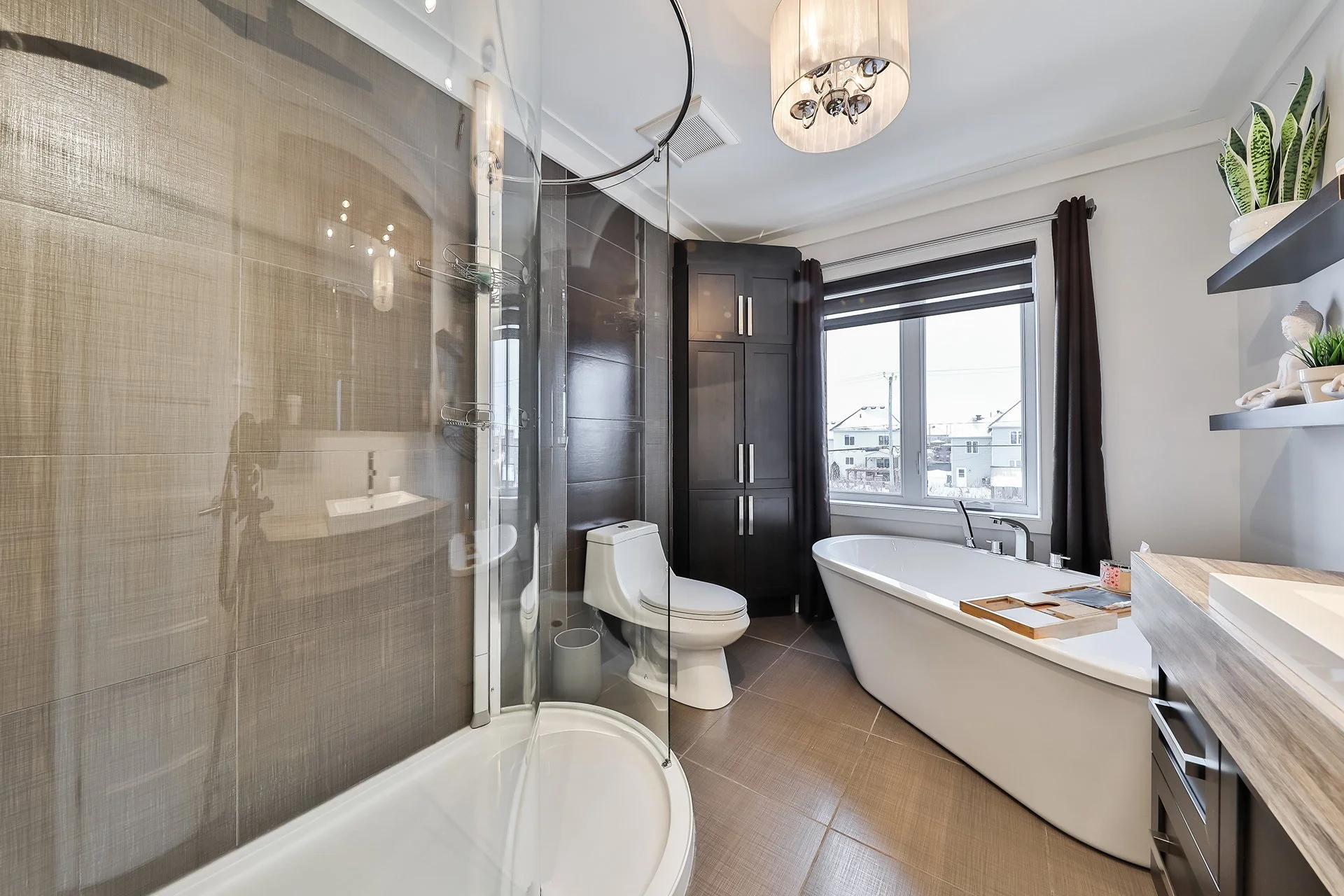 Une salle de bain moderne avec baignoire, toilettes, cabine de douche en verre, meuble en bois, étagères avec plantes et décorations, grande fenêtre laissant entrer la lumière naturelle.