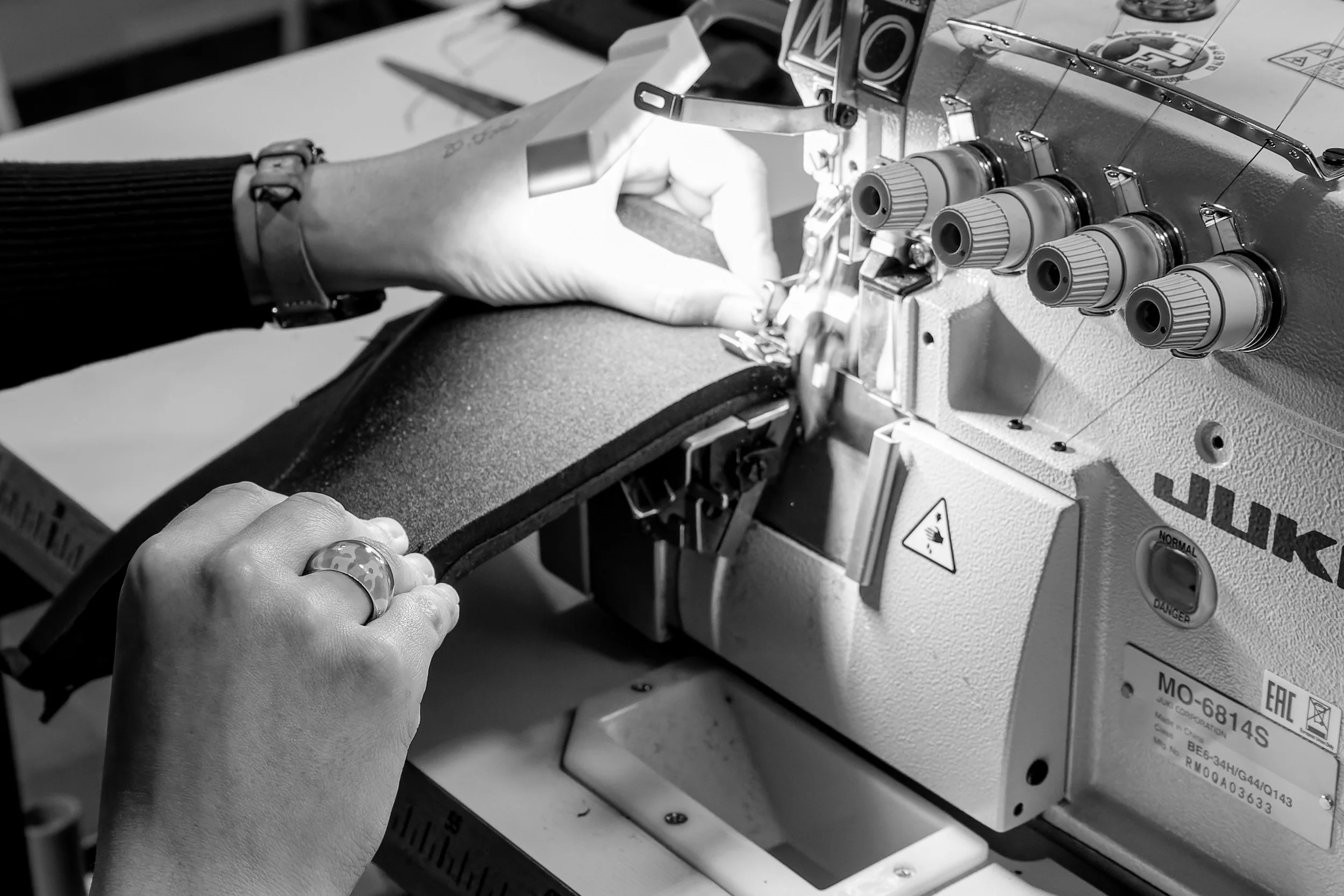 Deux mains manipulent une machine à coudre industrielle, utilisant un pointeur et un loquet pour ajuster la couture sur un tissu foncé.