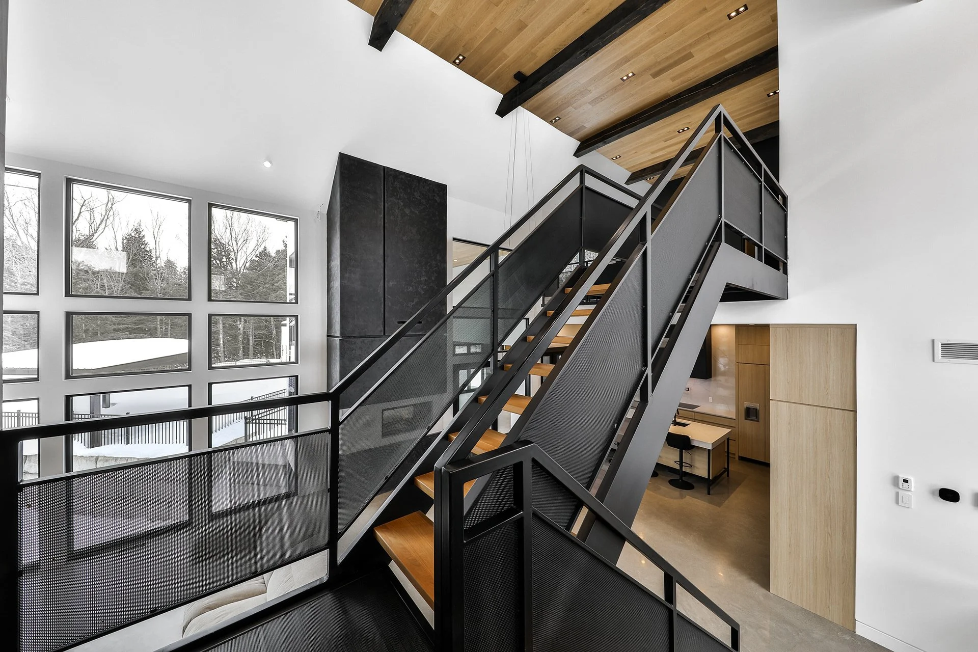 Escalier intérieur moderne avec rampe en métal et marches en bois, dans une maison contemporaine avec grandes fenêtres, plafond en bois et murs blancs.