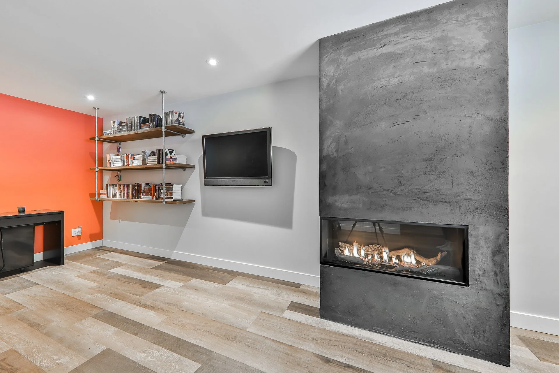 Salon avec cheminée moderne noire, télévision à l'écran plat, étagère en bois avec des livres, mur rouge-orange sur la gauche, mur blanc clair, sol en bois clair.