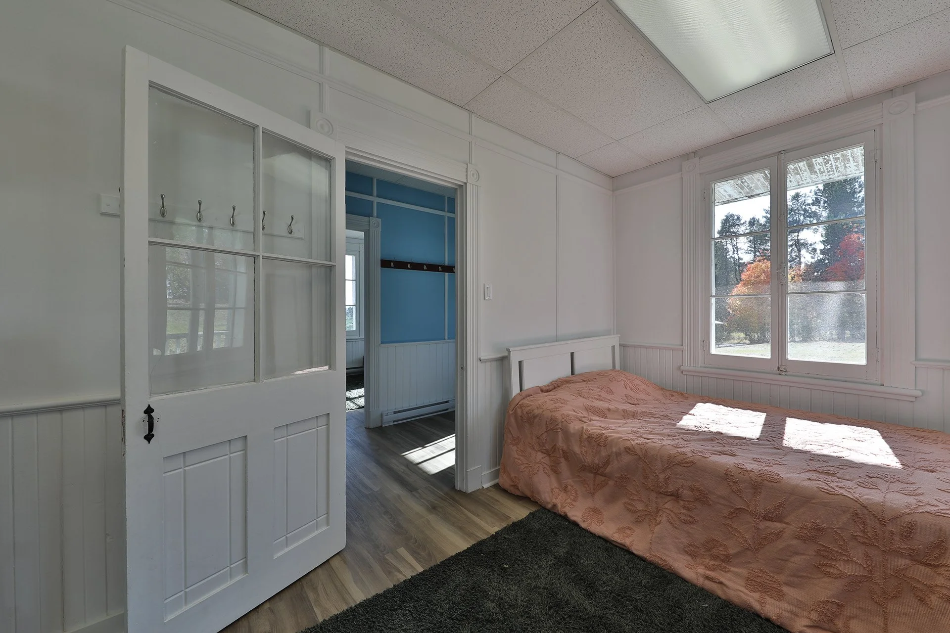 Chambre avec lit simple recouvert d'une couverture beige, une fenêtre laissant entrer la lumière, murs blancs, plancher en bois clair, porte ouverte vers un couloir ou une autre pièce avec murs bleus.