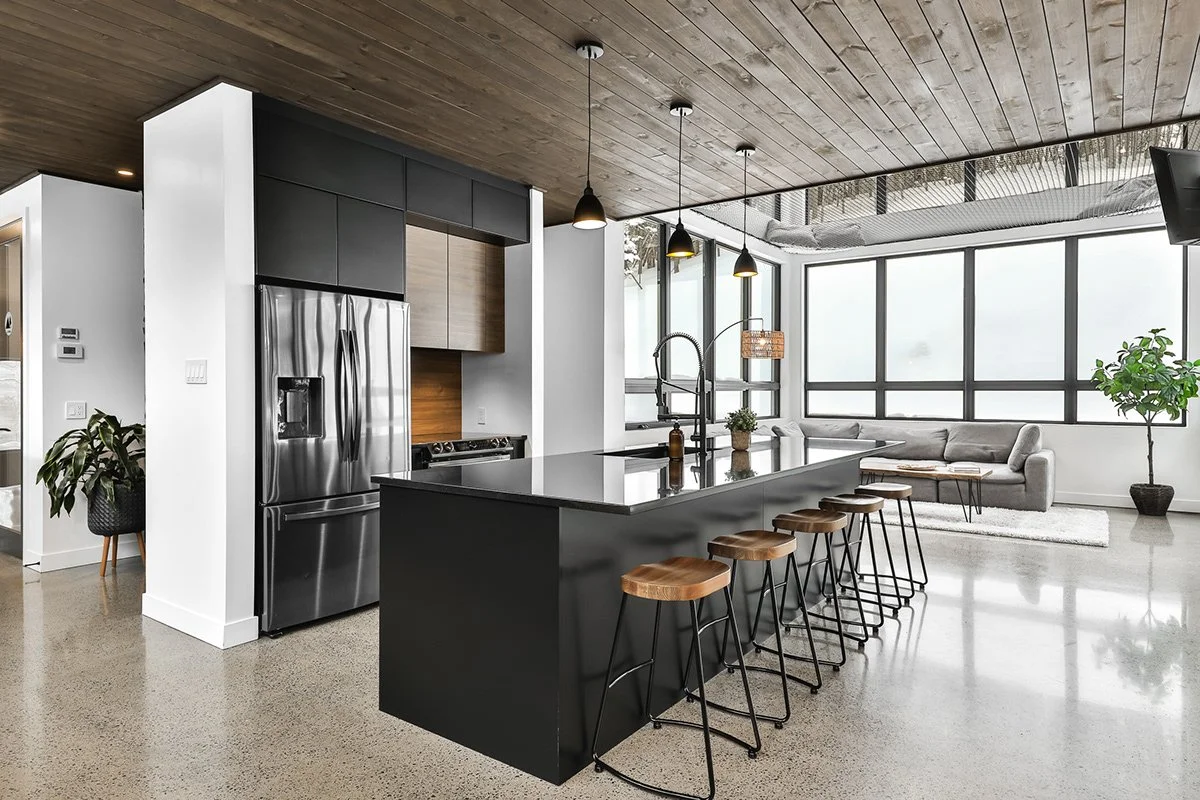 Cuisine moderne avec un grand îlot noir, des tabourets en bois, et un coin salon lumineux avec de grandes fenêtres et des plantes.