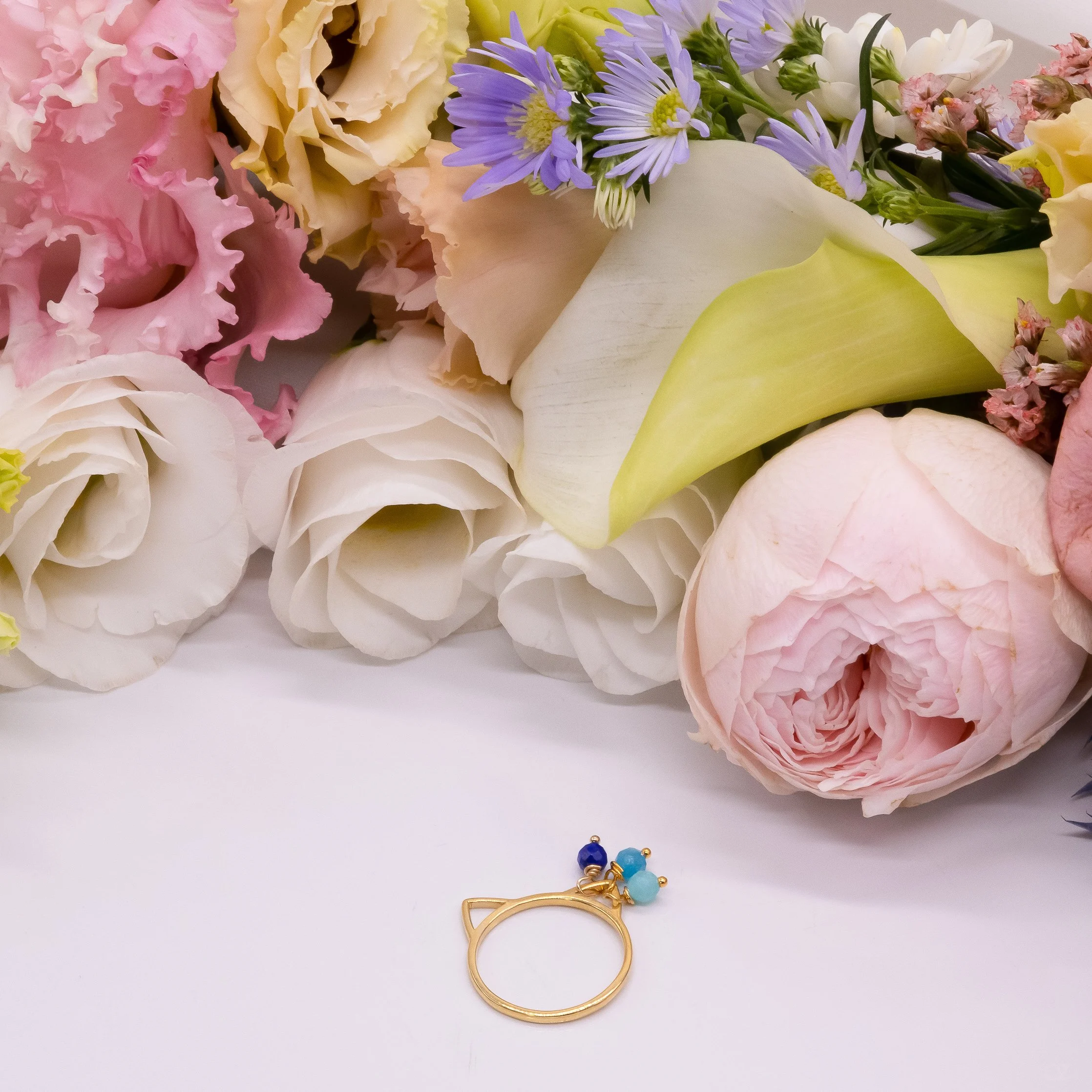 Un bouquet de fleurs avec des roses, des lis, et des fleurs violettes, ainsi qu'une bague dorée avec des perles bleues, posé sur une surface blanche.