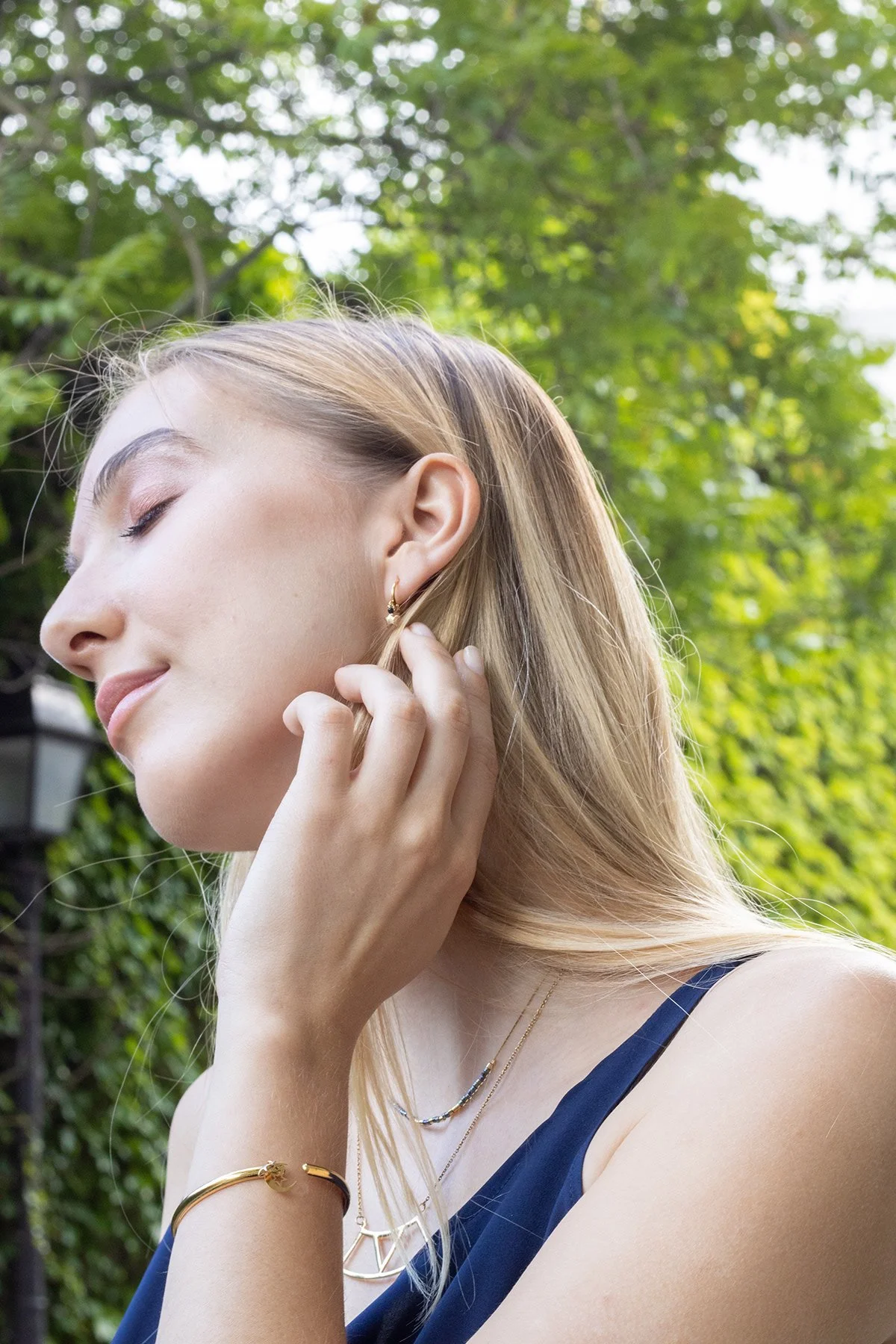 Jeune femme blonde avec des bijoux, les yeux fermés, touchant son oreille, en extérieur avec des arbres verts en arrière-plan.