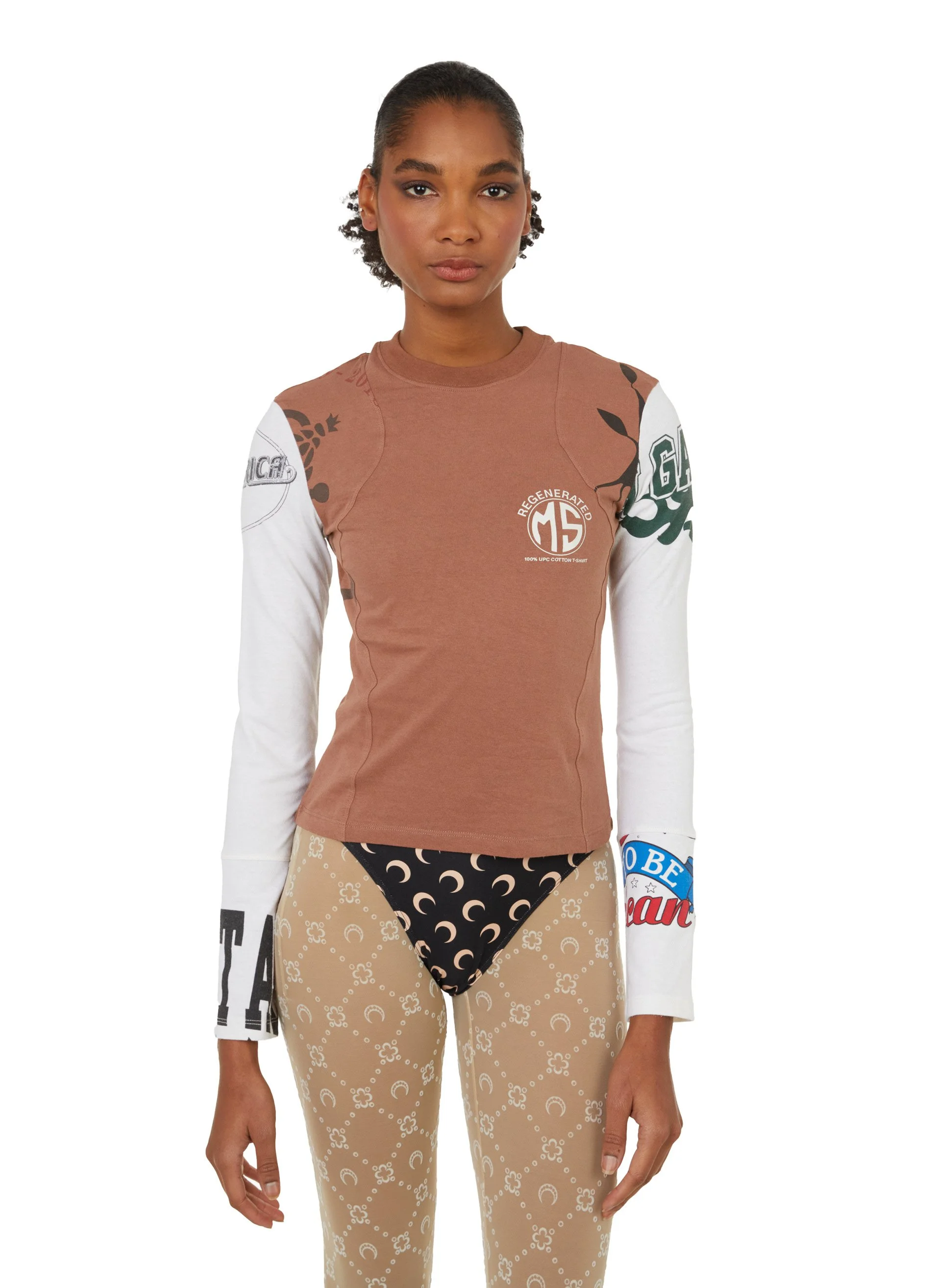 une femme debout contre un fond blanc, portant un t-shirt à manches longues avec des logos et des textes, et un pantalon beige avec un motif de lunes et de petits cercles.