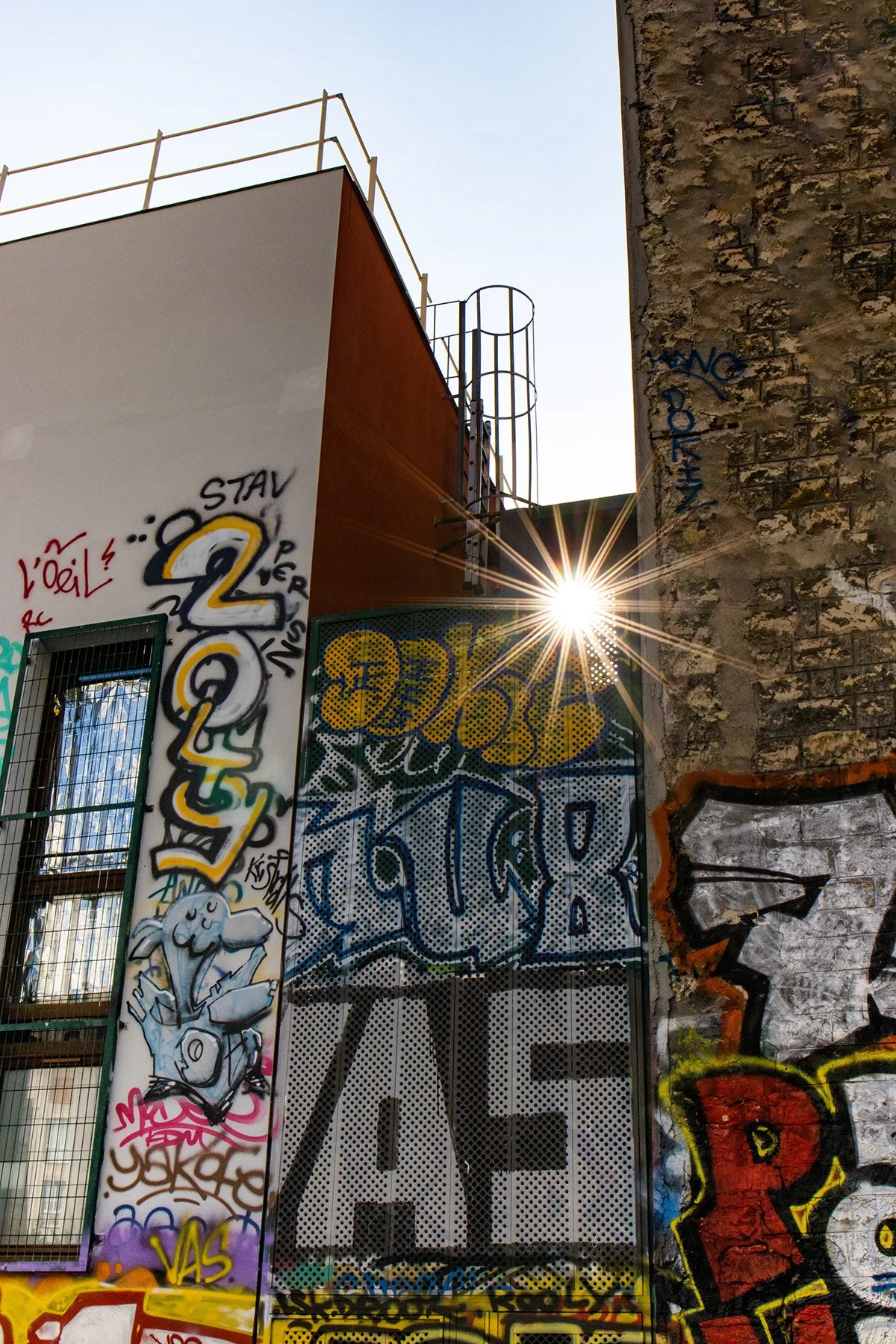 Murale urbaine avec graffiti coloré, mur en pierre, grille de fenêtre, soleil brillant derrière un bâtiment, et un panneau avec le mot 'LOVE' en lettres majuscules.
