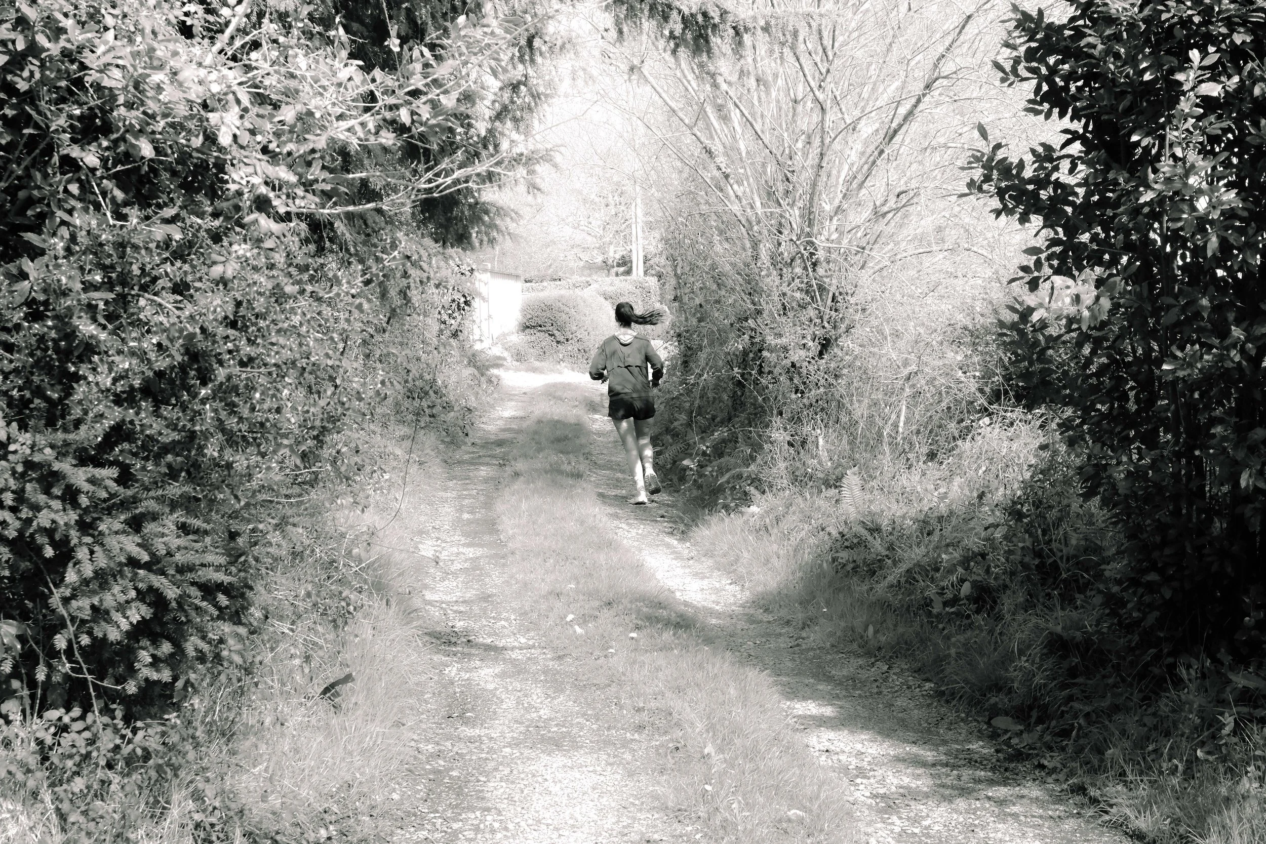 Une jeune fille court sur un sentier de campagne bordé d'arbustes et d'arbres sans feuilles, avec un ciel clair en arrière-plan.