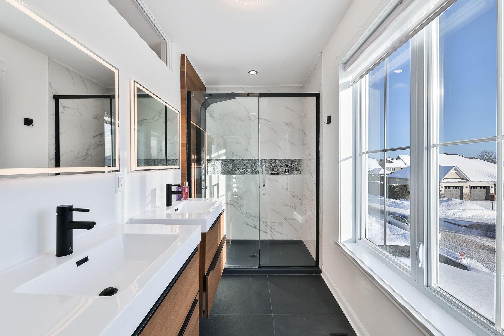 Une salle de bain moderne avec deux lavabos, un miroir grand, une cabine de douche en verre, et une grande fenêtre offrant une vue sur une scène enneigée extérieure.