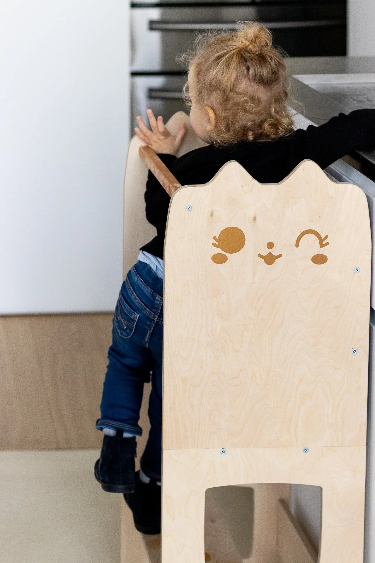 Enfant avec des cheveux bouclés roux assis sur une chaise en bois avec un visage d'animal souriant dessiné dessus, dans une cuisine.