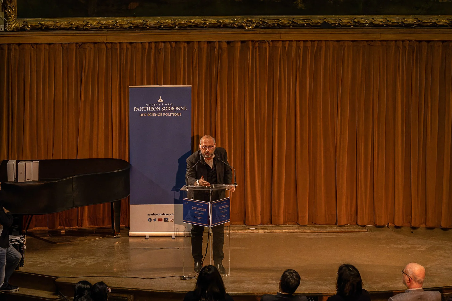 Un homme parle lors d'une conférence ou d'un discours devant un public dans une salle avec un rideau en bois derrière lui.