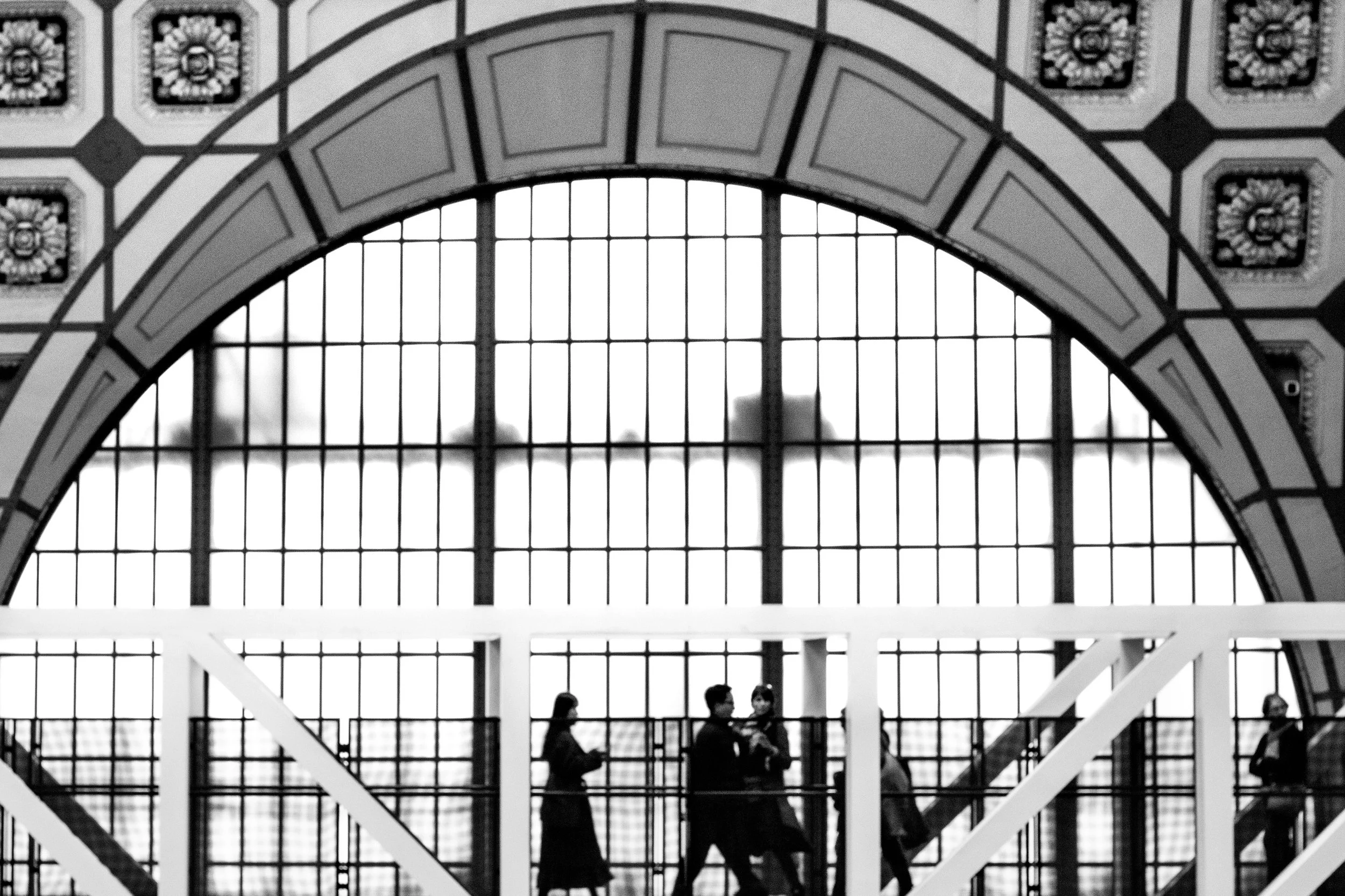 Vue en noir et blanc de personnes discutant à l'intérieur d'une structure architecturale avec une grande fenêtre de style industriel en arrière-plan, ornements décoratifs au plafond.