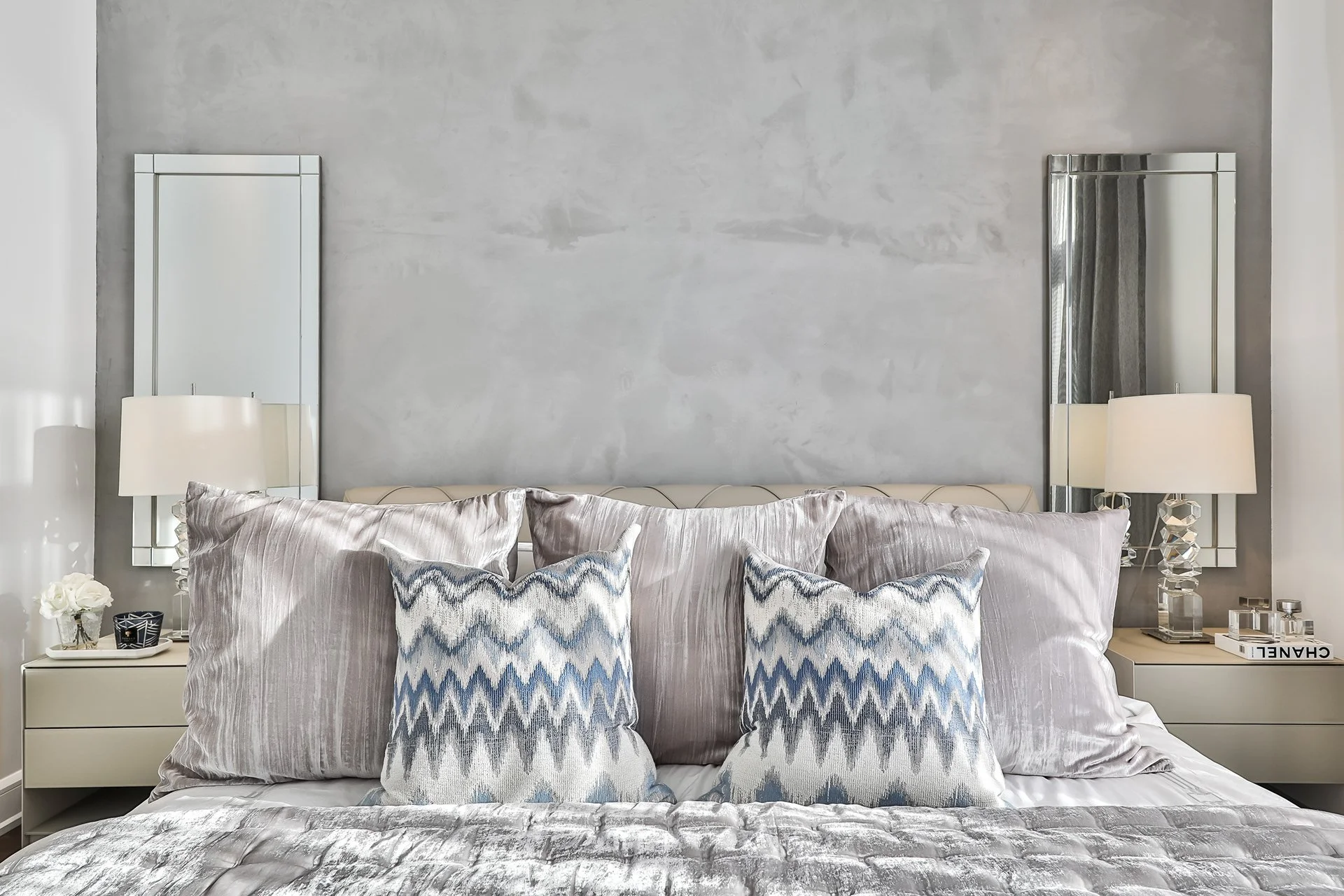 Lit avec plusieurs coussins gris et motifs bleus, deux tables de chevet avec lampes et décorations, murs gris en arrière-plan.