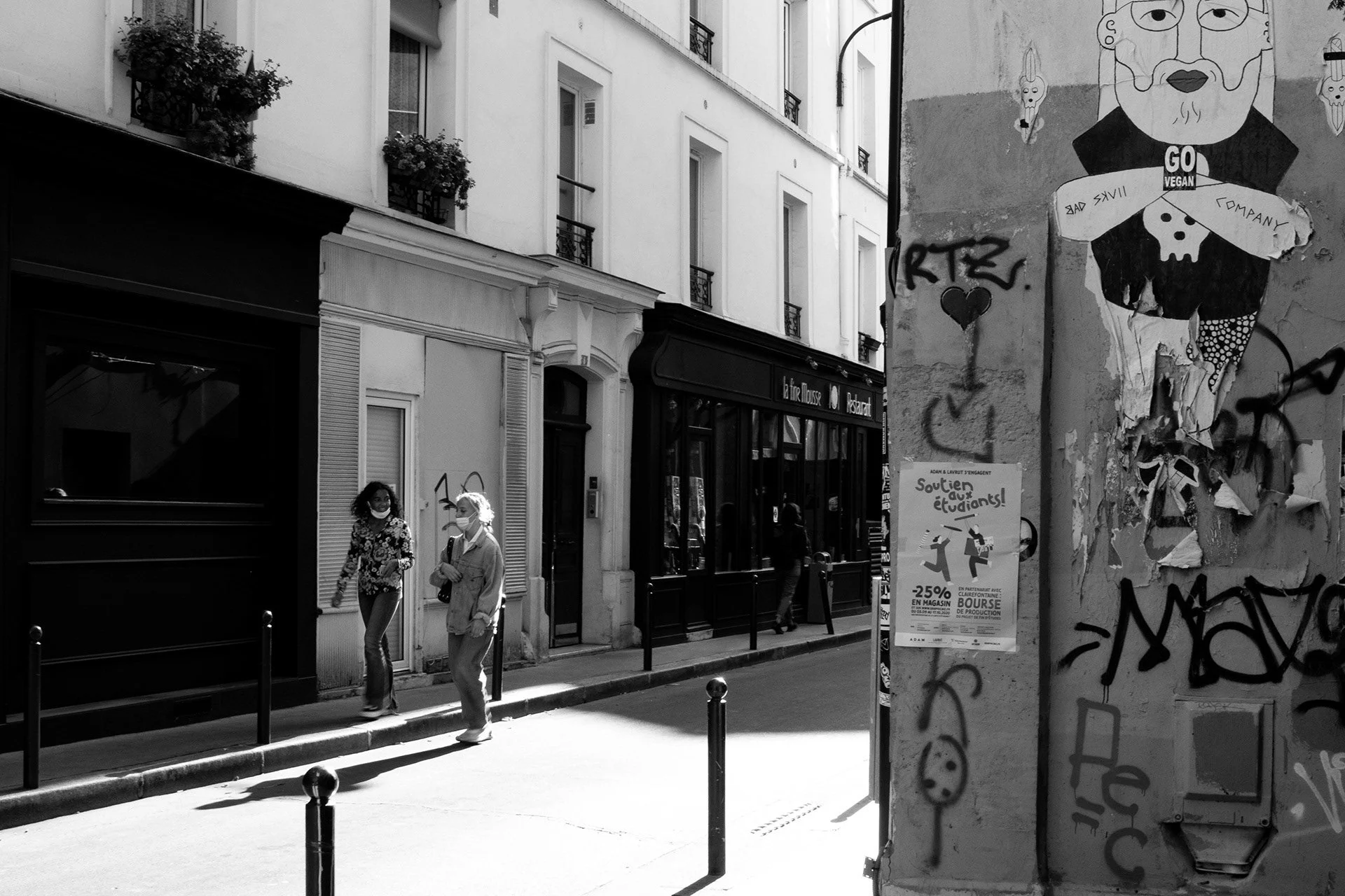 Rue urbaine avec deux personnes portant des masques, une boutique avec vitrine noire, graffitis et un poster à droite.