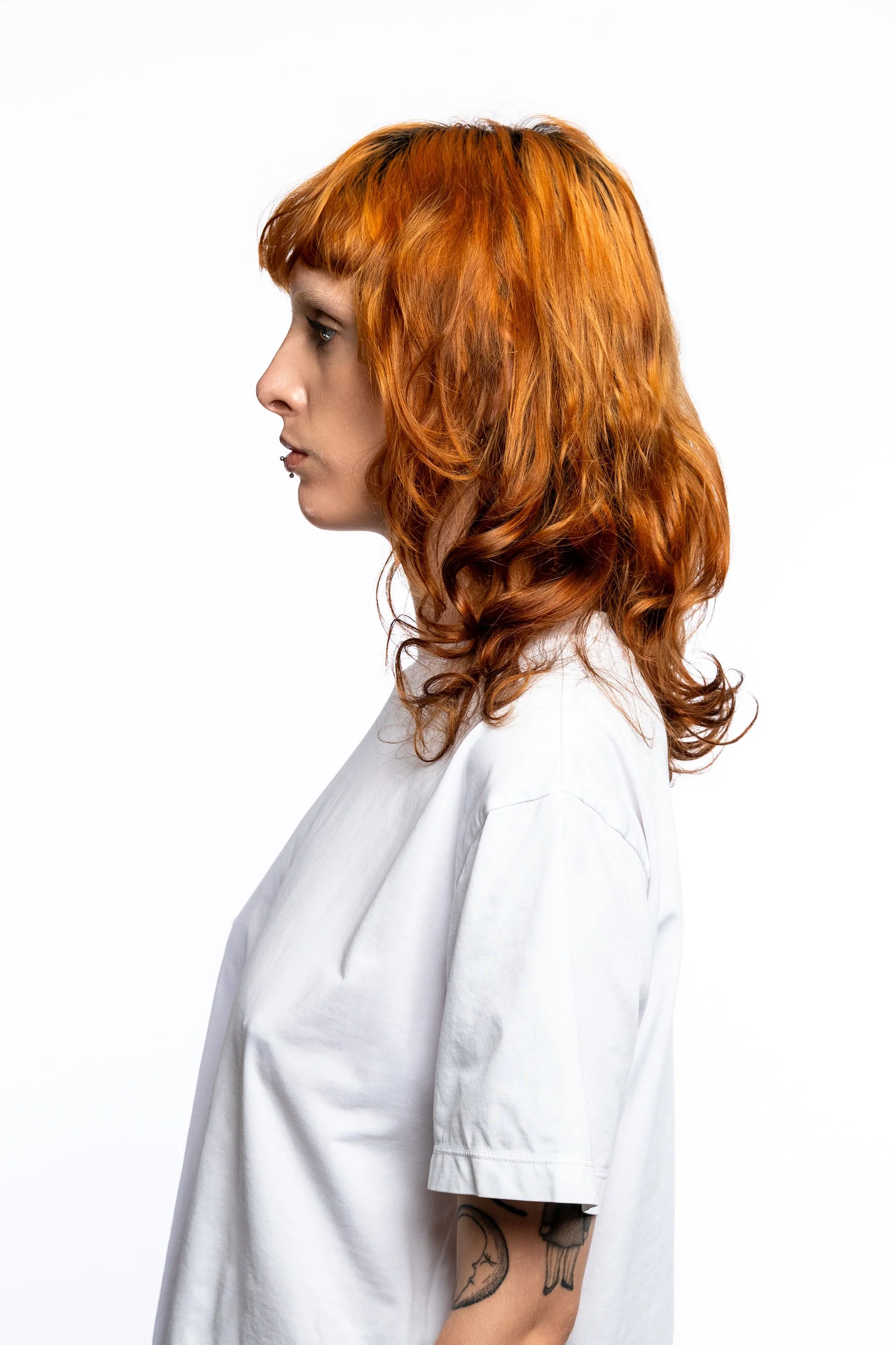 Femme aux cheveux roux et coupe en couches, portant un t-shirt blanc, vue de profil contre un fond blanc.