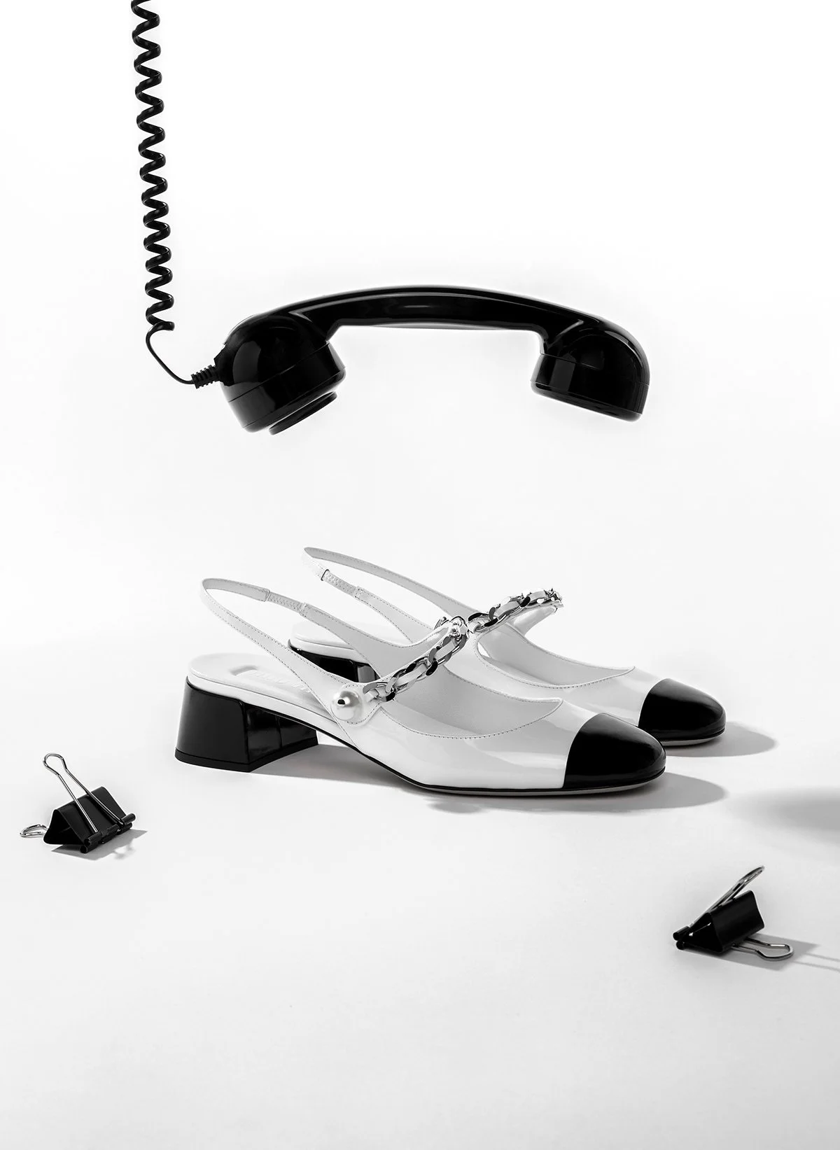 Des chaussures à talons blancs avec des bouts noirs et chaînes en métal, un téléphone fixe noir suspendu, et deux pinces en bas à gauche et à droite, tous disposés sur un fond blanc.