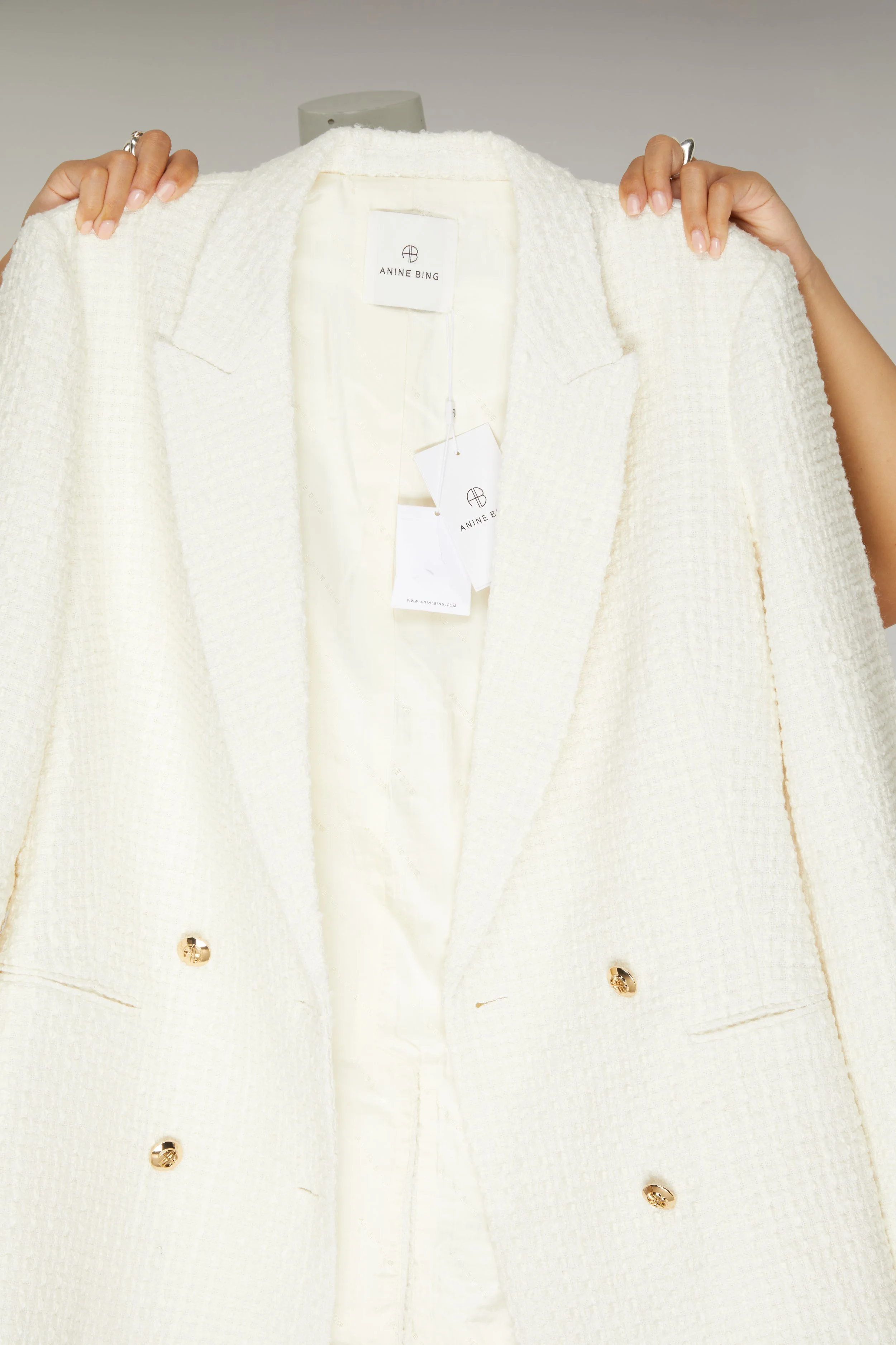 Une personne tient un blazer blanc avec des boutons dorés, portant une étiquette de marque 'Anine Bing'.