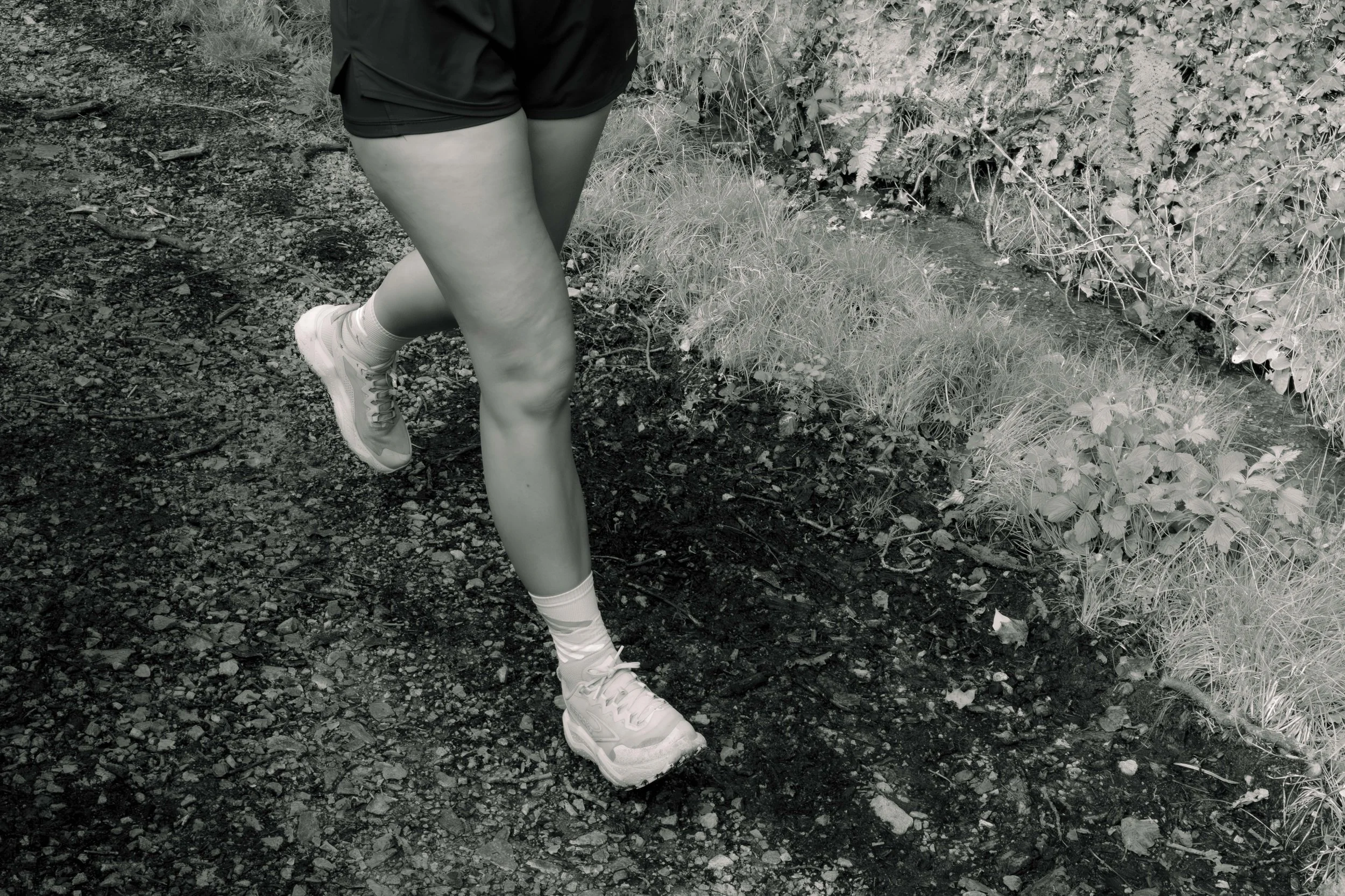 Une personne court ou marche dans un sentier en plein air, portant des chaussures de sport, des chaussettes blanches, un short noir et un haut noir, avec de la végétation luxuriante sur le côté.