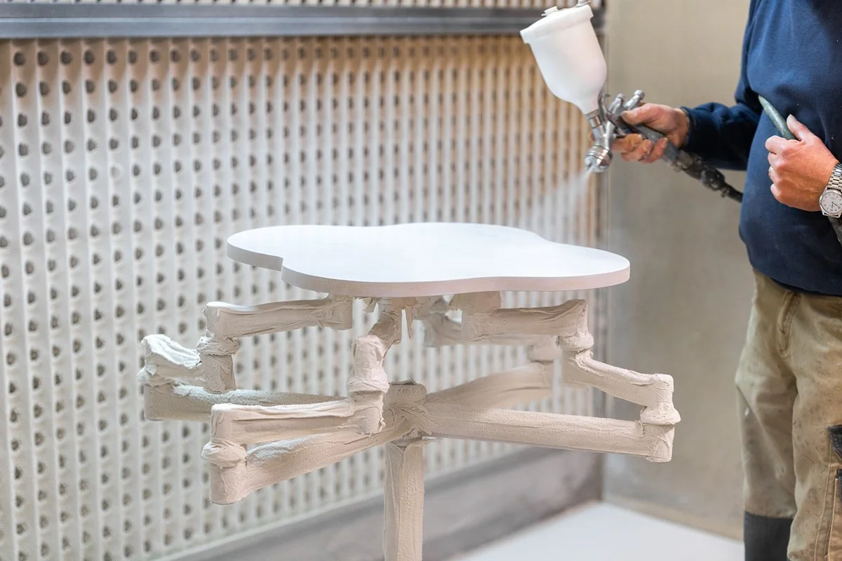 Un artisan utilisant un pistolet à peinture pour peindre un meuble en bois blanc. Le meuble est en cours de fabrication ou de finition.