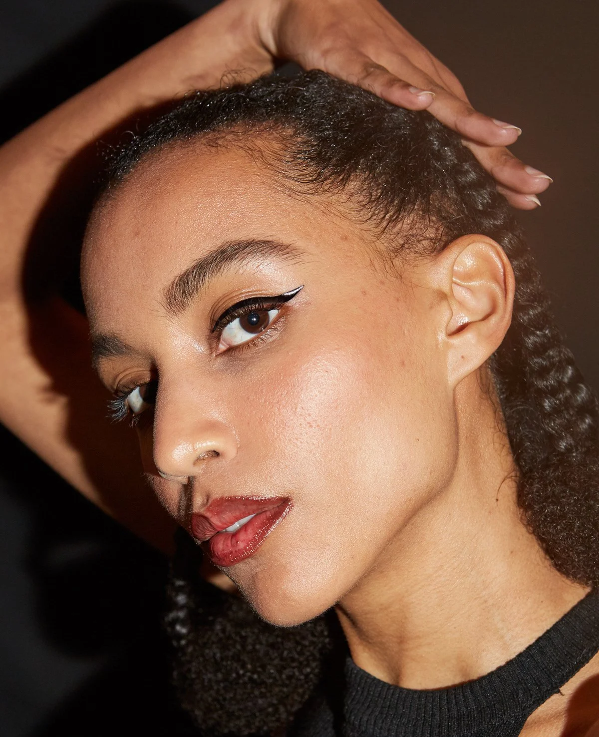 Portrait d'une femme avec maquillage sophistiqué et cheveux curls, portant un haut noir, dans un éclairage chaud.