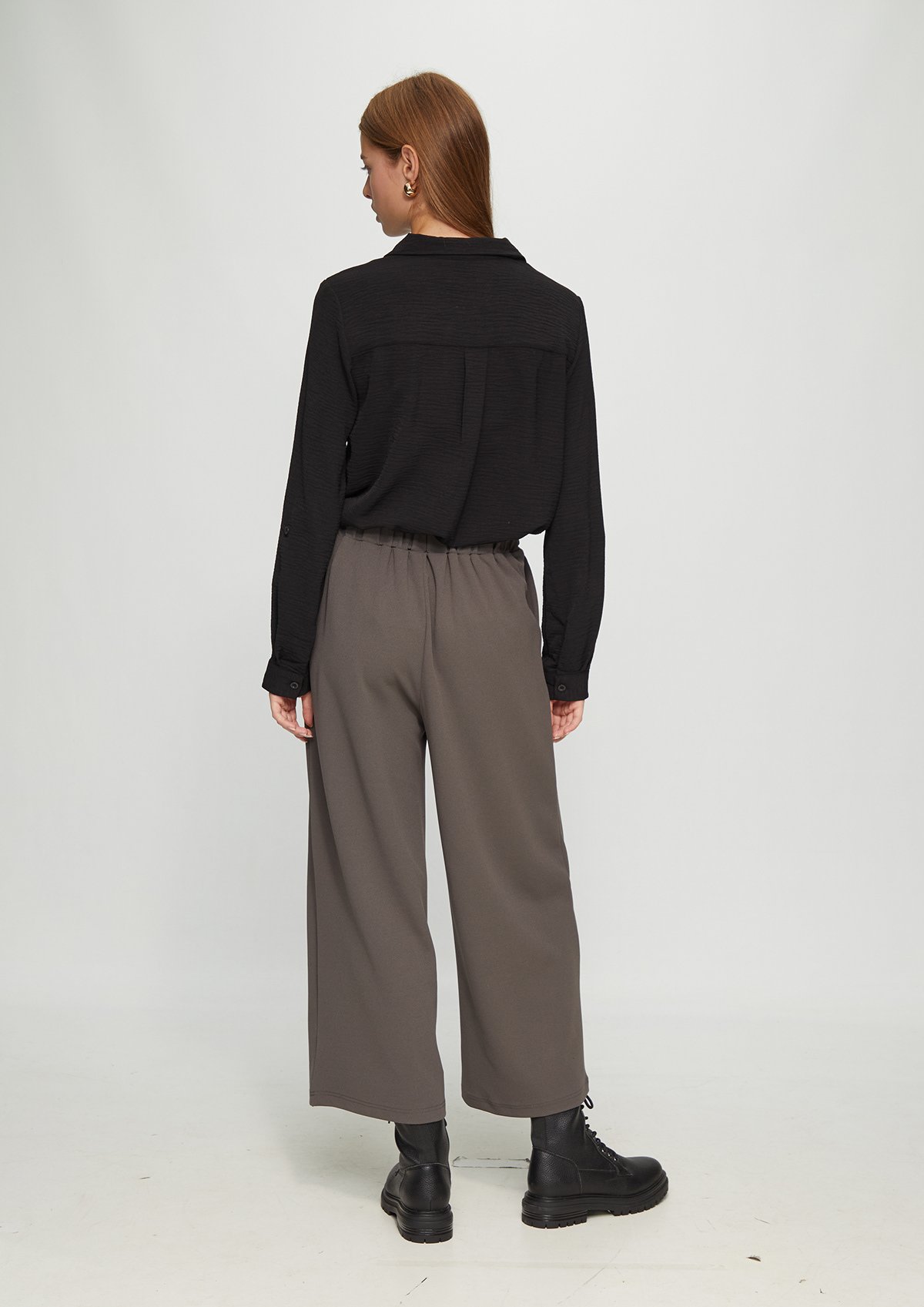 Femme portant une chemise noire, un pantalon gris ample et des bottes en cuir noir, vue de dos.