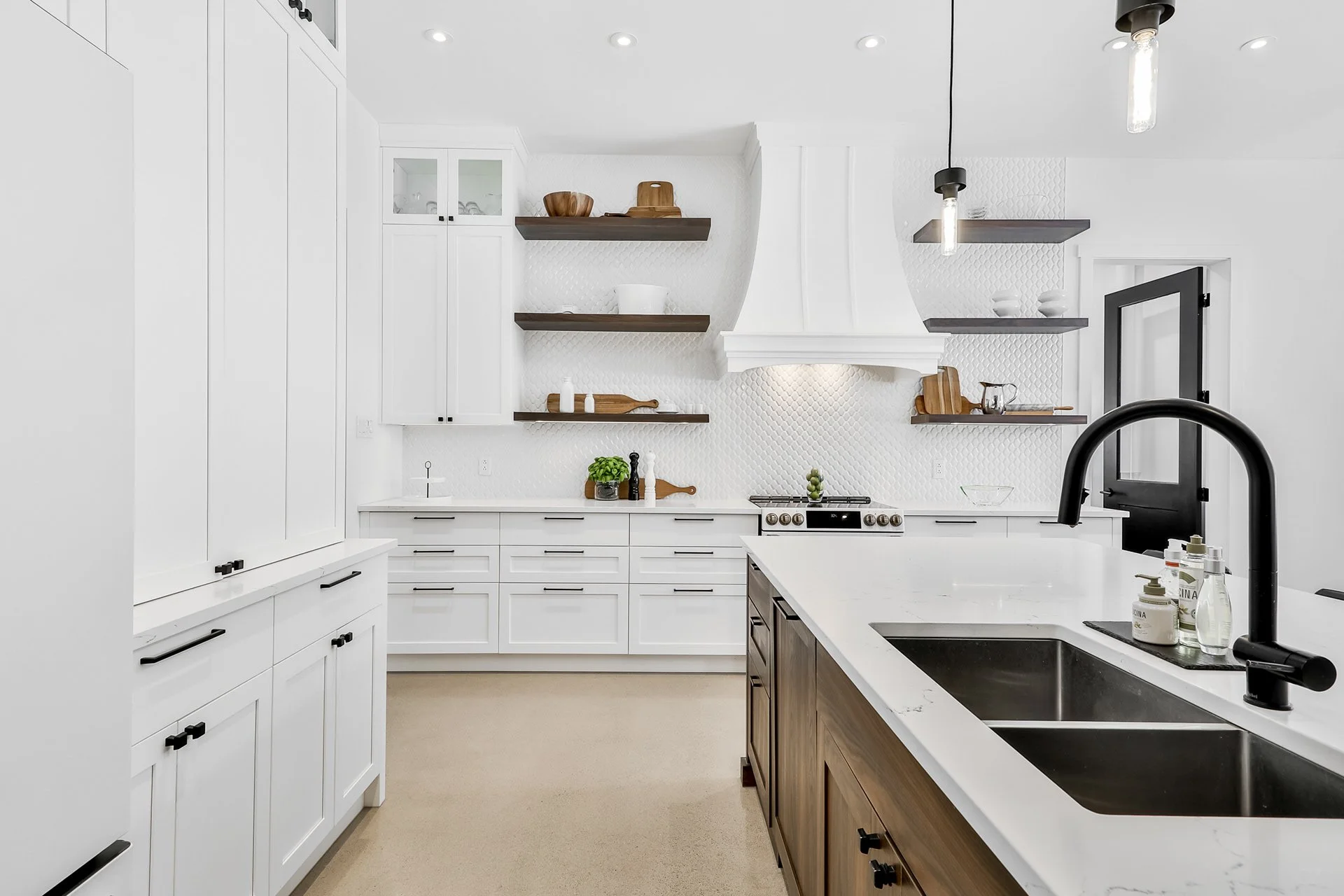 Cuisine moderne avec comptoir en marbre blanc, armoires blanches, étagères en bois, évier noir, et poutre de cuisson blanc, éclairage suspendu.