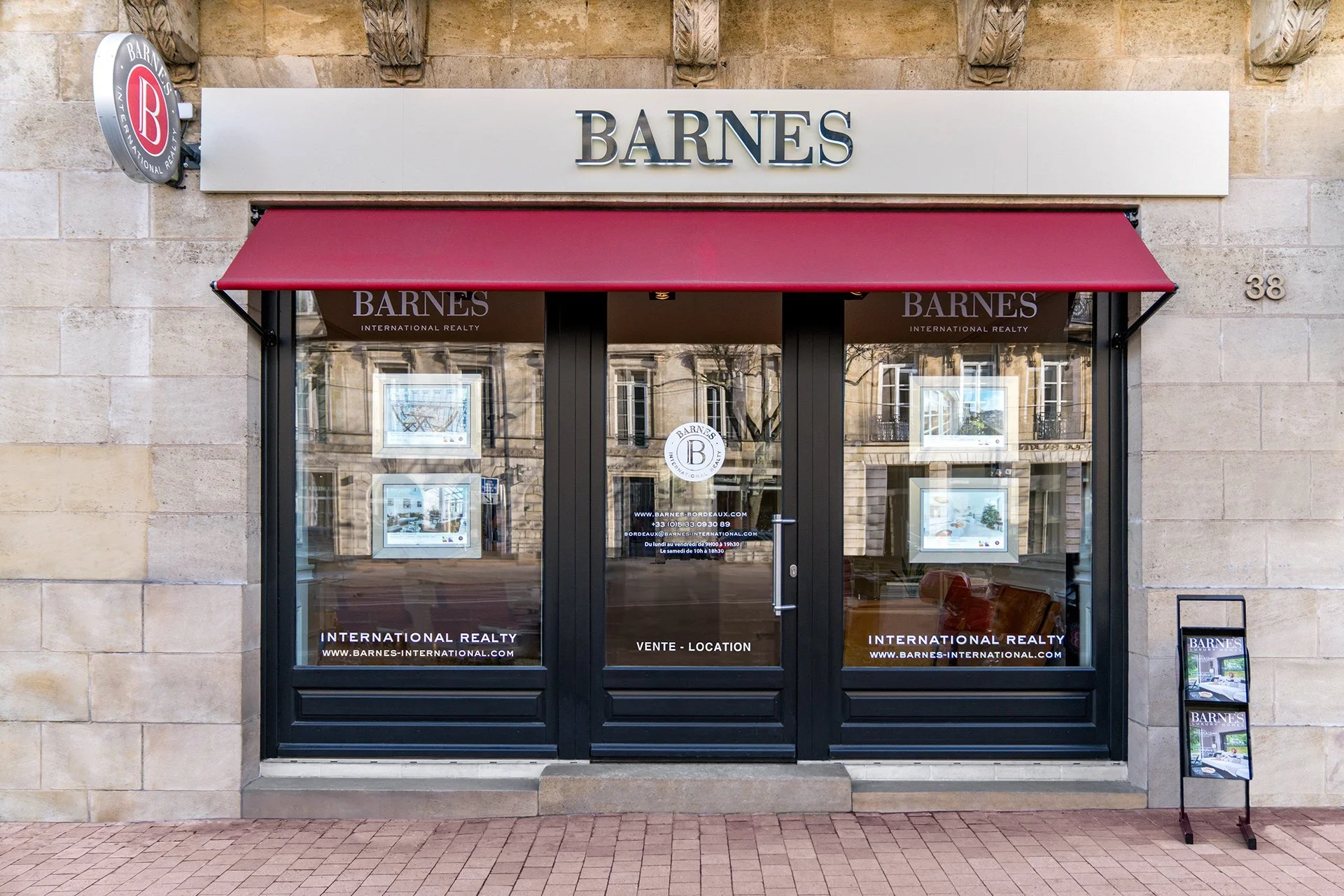 Vitrine d'agence immobilière avec le nom 'BARNES' en lettres noires sur un fond blanc, devant une façade de bâtiment en pierre, avec une enseigne ronde en haut à gauche et un panneau d'affichage devant la vitrine.
