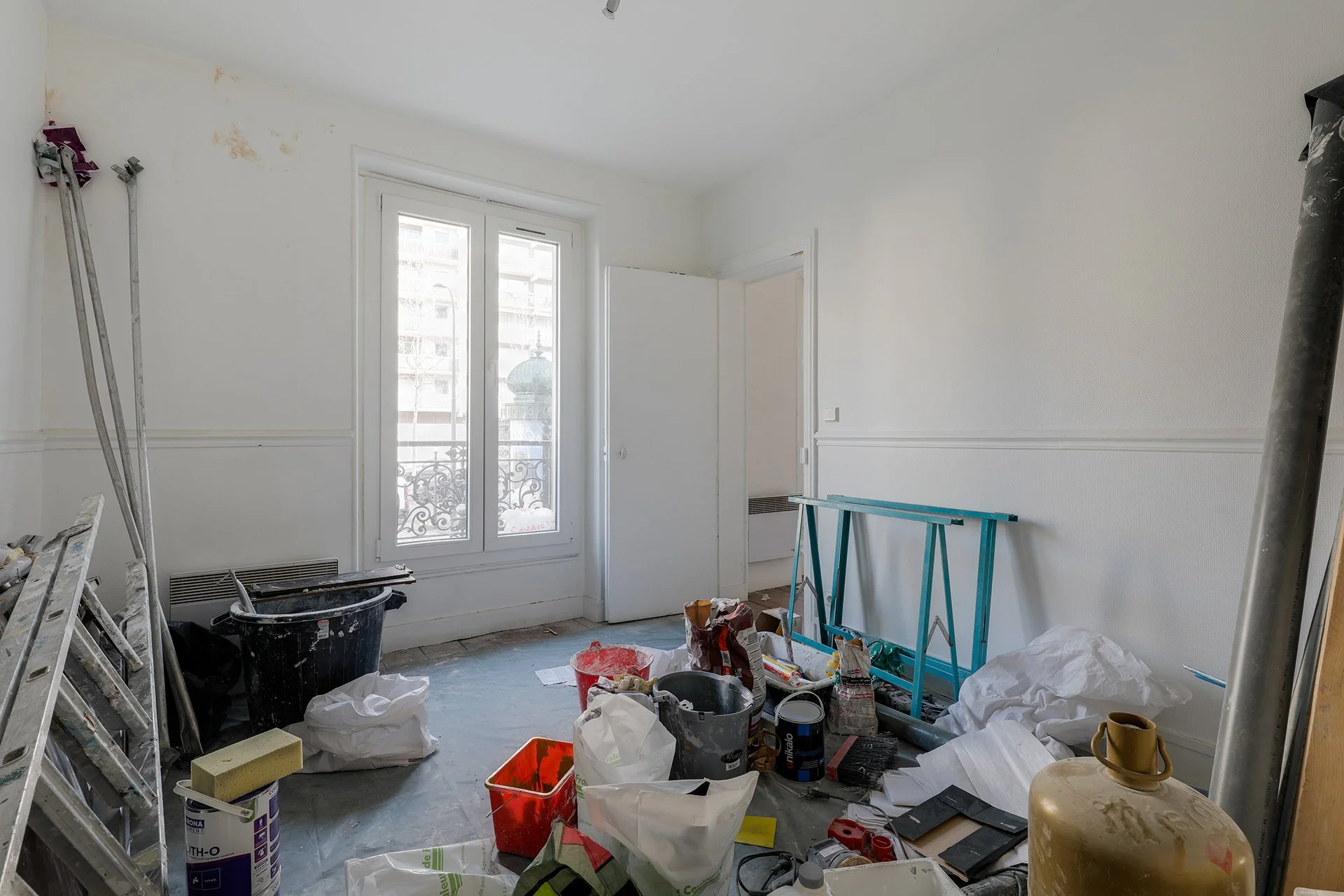 Une pièce en cours de rénovation avec des outils, des peintures, et des matériaux de construction éparpillés sur le sol, une fenêtre laissant entrer la lumière naturelle, et des murs en cours de peinture.