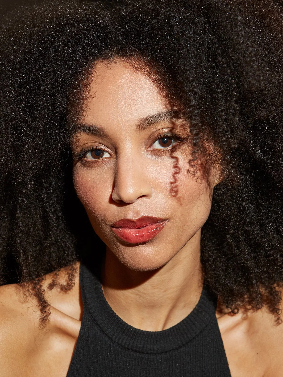 Portrait d'une femme avec de grands cheveux bouclés noirs, peau claire, souriant légèrement, portant un haut noir.
