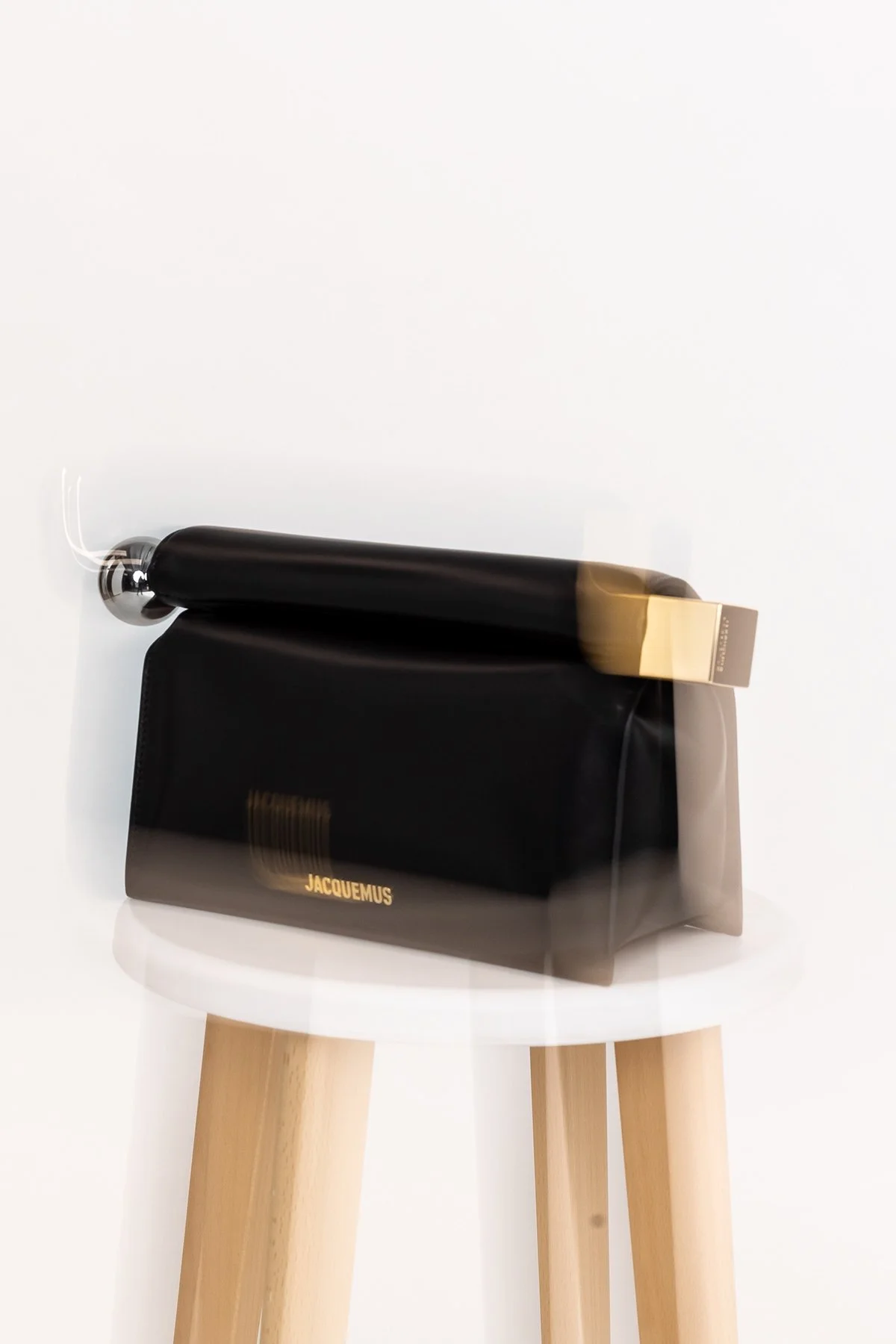 Une sac à main noir avec le mot 'JACQUEMUS' en or est placé sur un tabouret en bois clair, avec un rouleau à pâtisserie noir et or posé dessus, tout contre un mur blanc.