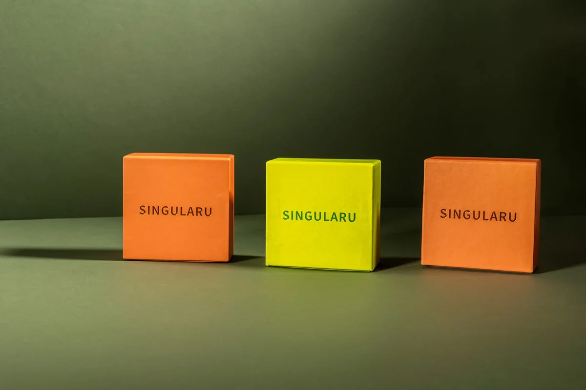 Trois boîtes colorées avec le mot 'SINGULARU' écrit dessus, alignées sur une surface verte, avec un fond vert flou.