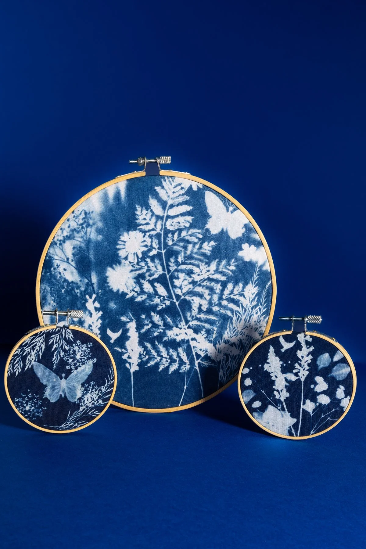 Trois cerceaux de broderie avec des motifs identiques de feuilles et de papillons, disposés sur un fond bleu, avec une broderie en blanc sur un fond bleu.