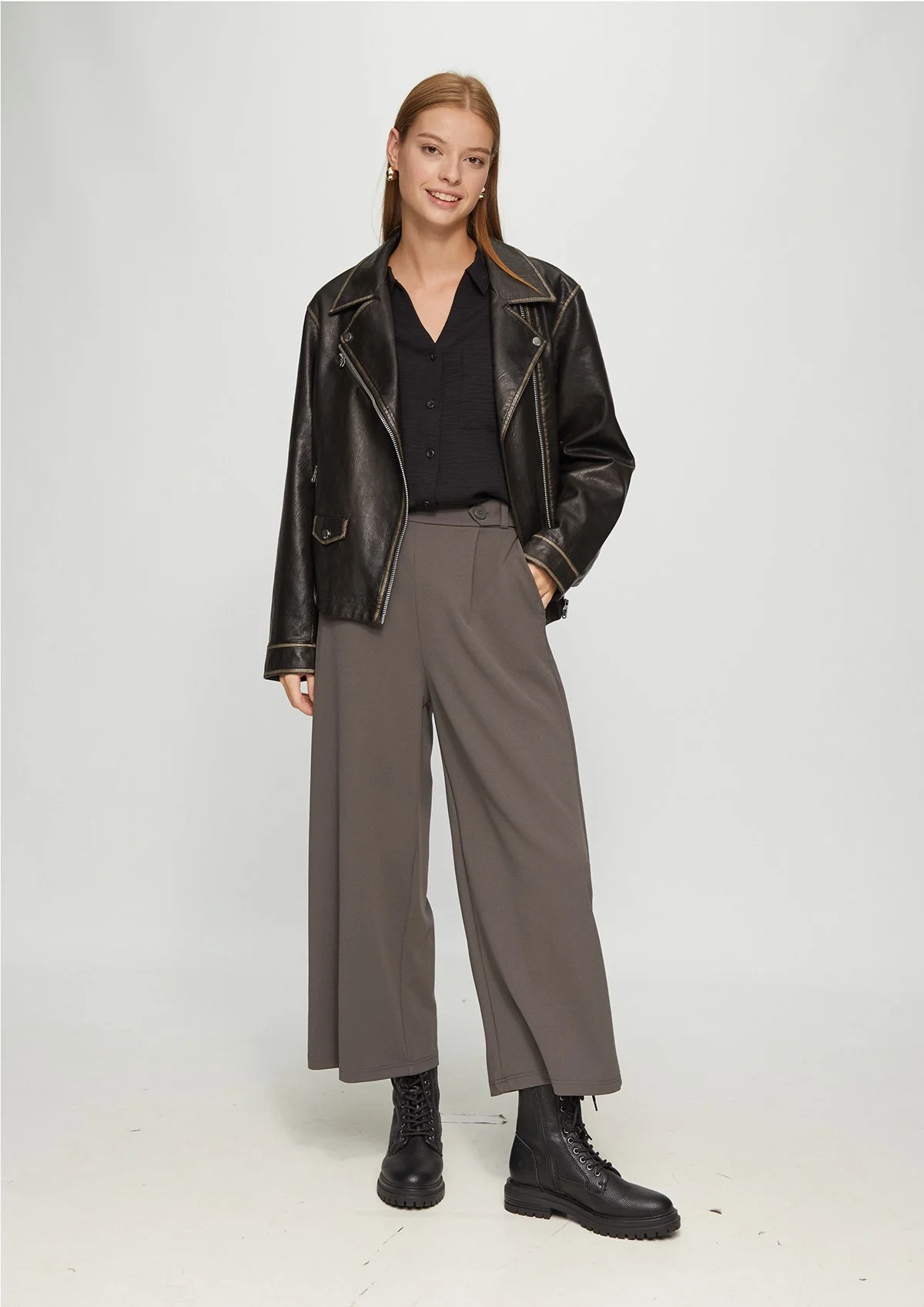 Une femme souriante portant un blouson en cuir noir, un chemisier noir, un pantalon large gris et des bottes noires.