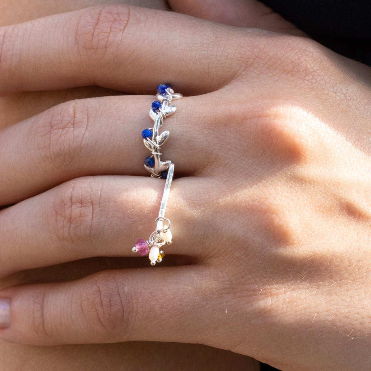 Une main porte une bague argentée ornée de petites pierres bleues et un bracelet assorti avec des pendentifs colorés, capturés sous une lumière naturelle.